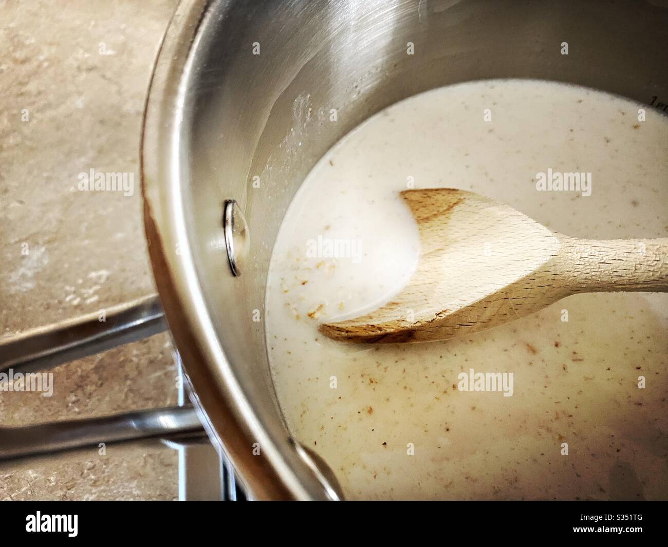 Ein Blick auf die Haferbrei, die auf einem Gasherd kochen. Köcheln Sie gerollte Hafer und Milch in einer silbernen Pfanne mit Milch. Gesundes Frühstück mit Holzlöffel. Stockfoto