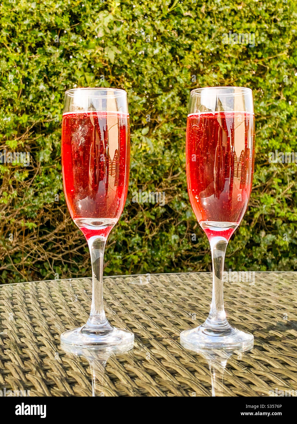 Zwei Flötengläser aus rosa Champagner auf der Glasplatte eines Tisches in einem Garten an einem Sommerabend Stockfoto
