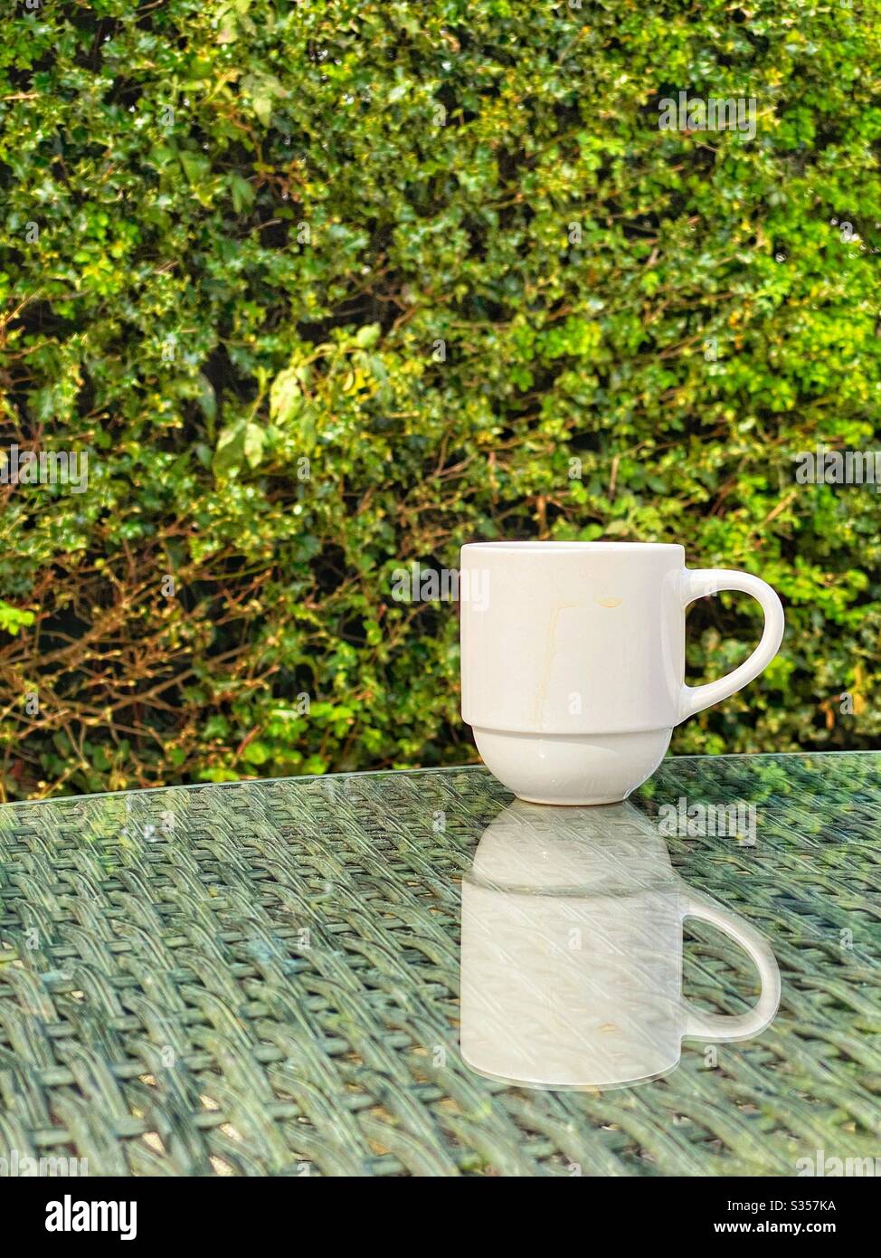 Weiße Kaffeetasse auf Gartenmöbel mit Spiegelung in Glas-Tischplatte Stockfoto