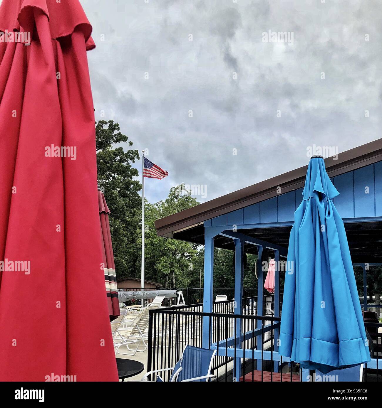 Poolbereich, sehr bewölkt, amerikanische Flagge, Bäume, Poolmöbel, rote, blau gefaltete Sonnenschirme, Pavillon, keine Menschen, Wetter, Kopierplatz Stockfoto