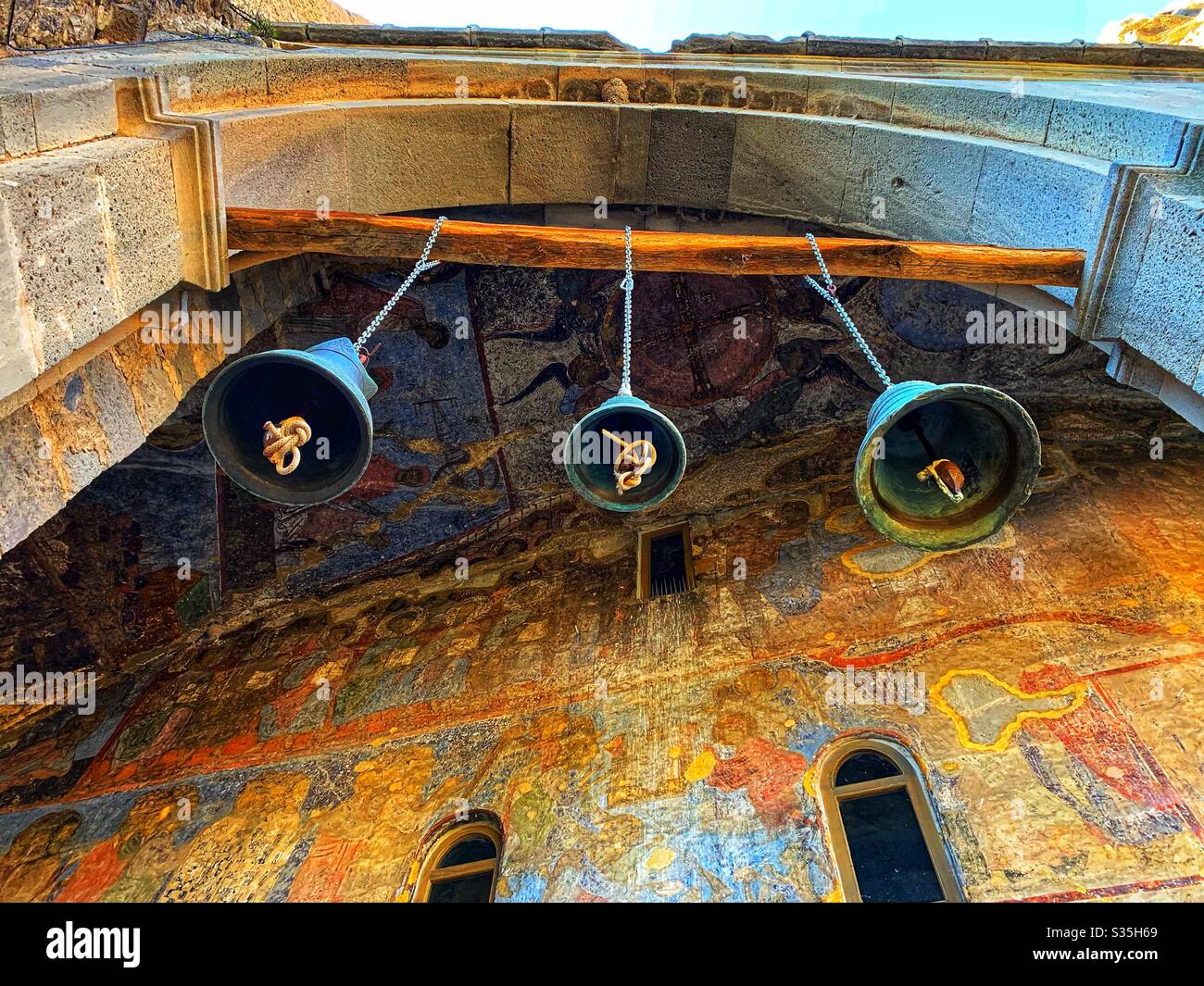 Drei Glocken, die von einem Holzbalken, gestützt von einem Bogen, mit einem Hintergrund der historischen religiösen Kunst aus dem 13. Jahrhundert in Vardzia in Georgien aufgehängt. - Smartphone-aufgenommenes Stockfoto