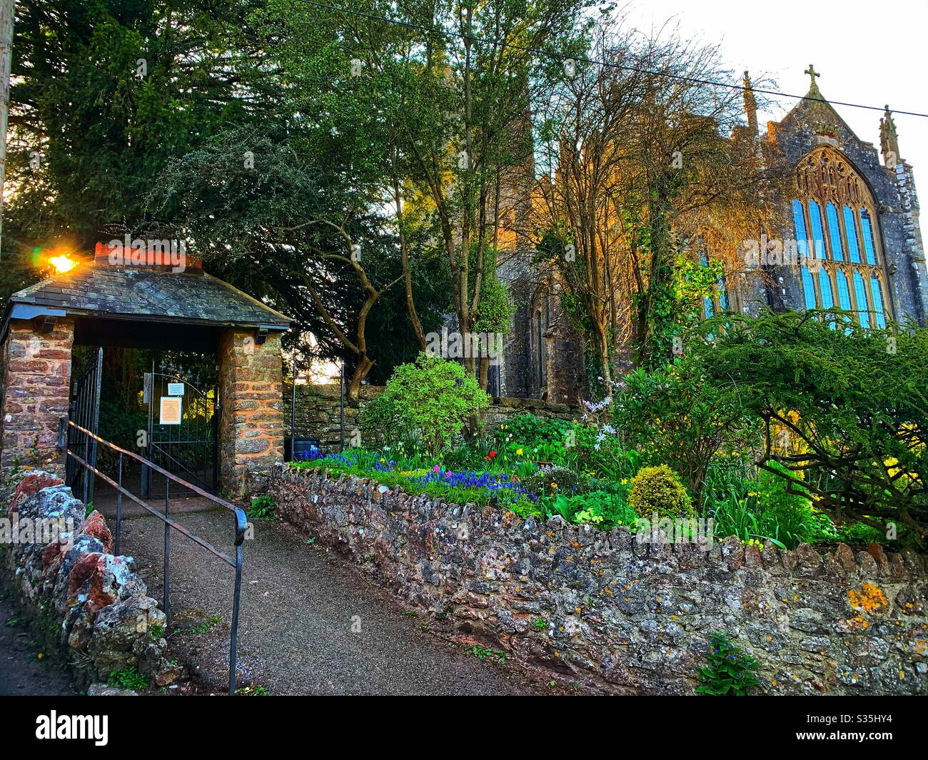 Eine Devon Village Kirche gegen Ende des Tages mit der untergehenden Sonne, Bäumen und Blumen. - Smartphone-aufgenommenes Stockfoto