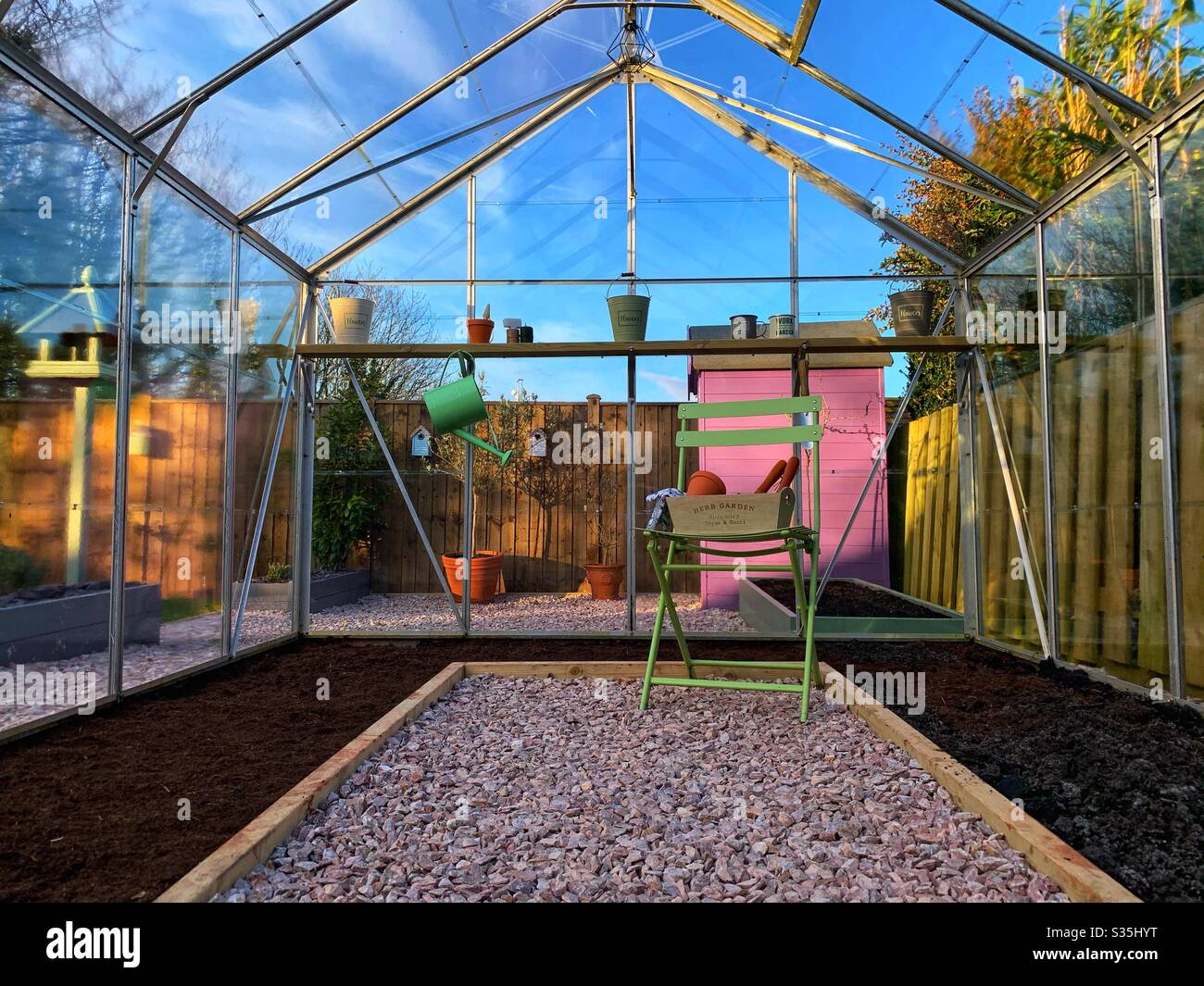 Ein Gewächshaus, bereit für die Pflanzung in. Ein grüner Metallstuhl mit Gartengeräten und einem Vogeltisch. Stockfoto