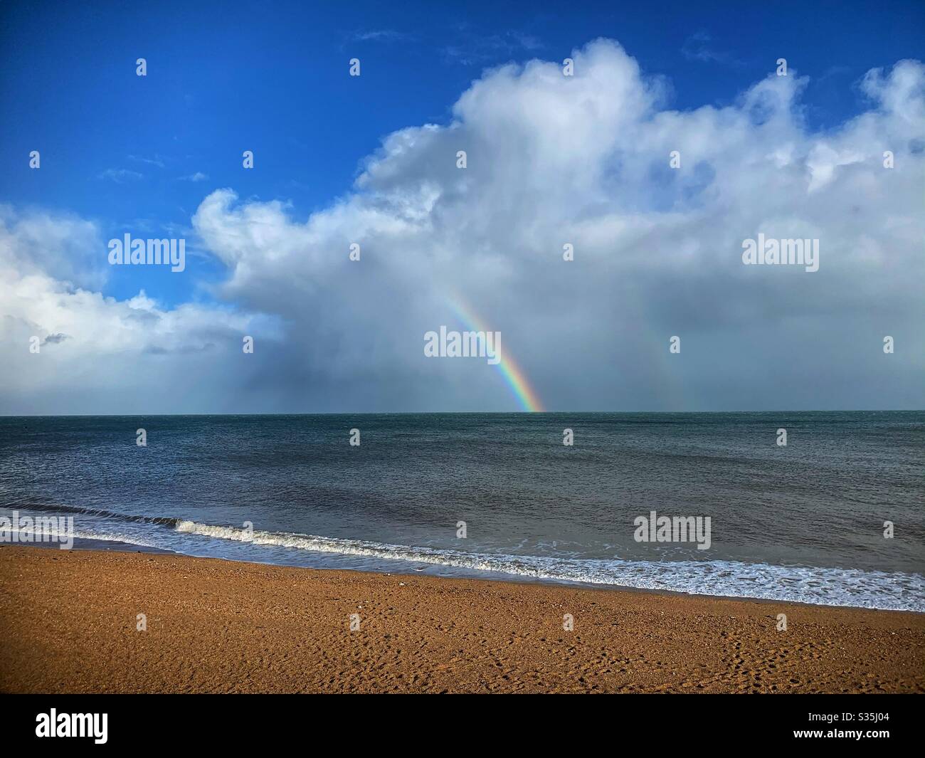 Regenbogen über dem Strand. Blauer Himmel mit weißen Wolken. - Smartphone-aufgenommenes Stockfoto