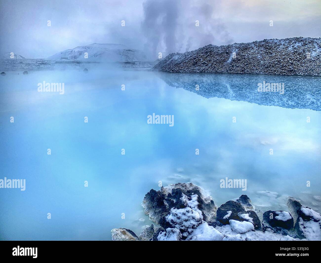 Dampf steigt aus einem blauen geothermischen Pool in Island. Umgeben von vulkanischem Gestein, bedeckt mit Schnee. - Smartphone-aufgenommenes Stockfoto