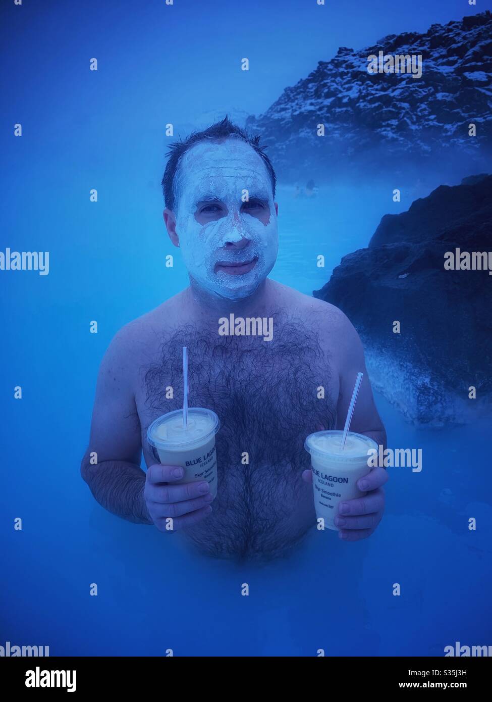 Mann, der in einem geothermischen Pool an der Blauen Lagune in Island steht, während er eine Kieselsäure-Gesichtsmaske trägt und zwei Bananenmilchshakes hält. Umgeben von milchig blauem Wasser und vulkanischem Gestein. Stockfoto