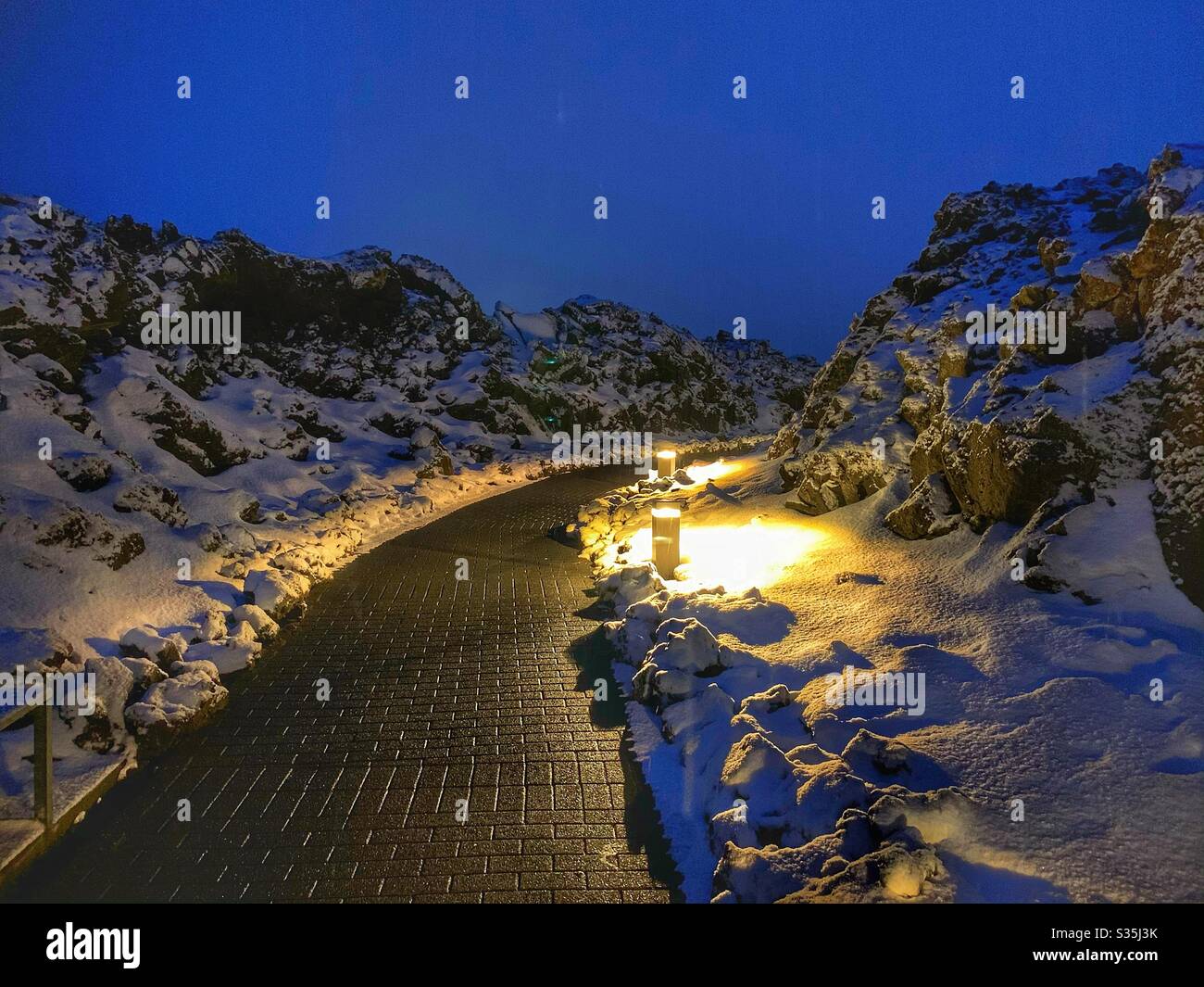 Pfad umgeben von schwarzem Vulkangestein, bedeckt mit Schnee und beleuchtet durch Gehwege Beleuchtung in blauer Stunde. Eintritt zur Blauen Lagune, Island. - Smartphone-aufgenommenes Stockfoto