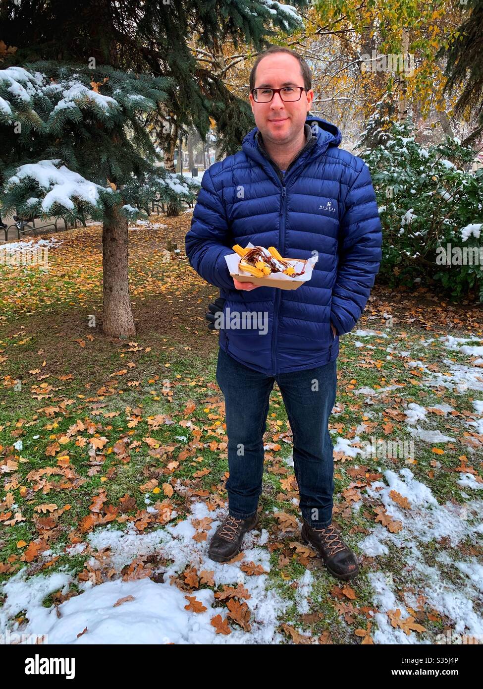 Mann hielt ein Tablett mit frisch zubereiteten Churros, die mit Schokoladensauce bedeckt waren, während er in einem verschneiten Park stand, der von Bäumen umgeben war. - Smartphone-aufgenommenes Stockfoto