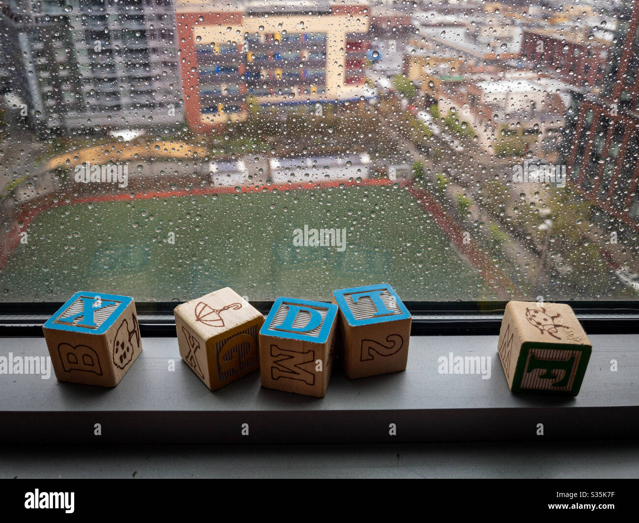 Buchstabenwürfel auf dem Rahmen eines Fensters, das an einem regnerischen Tag über ein leeres Sportfeld blickt - Smartphone-aufgenommenes Stockfoto