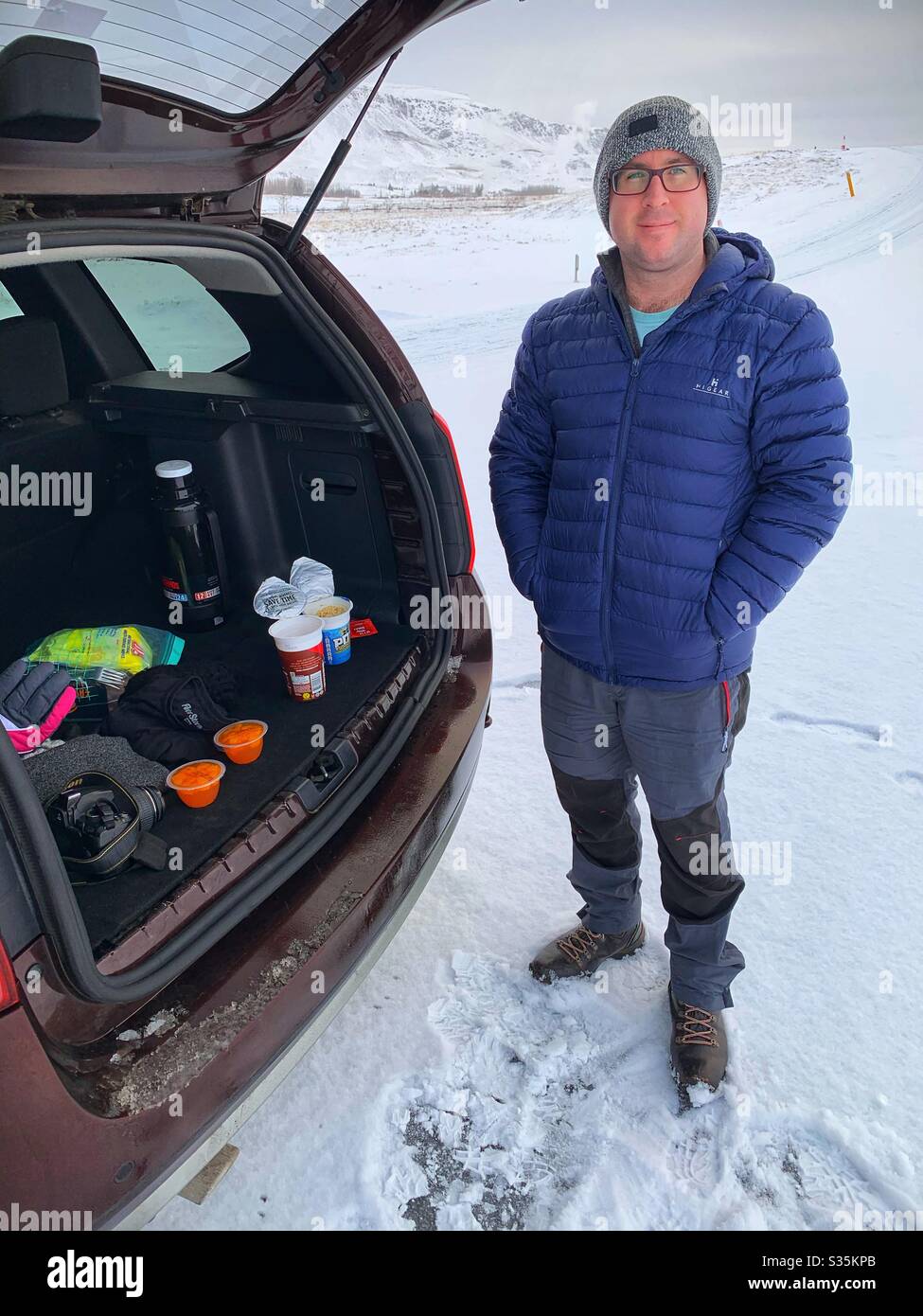 Mann in Outdoor-Winterkleidung, der hinter einem Auto steht und die Kofferraumtür nach oben zeigt, und ein warmes Mittagessen mit einer Thermoskanne im Schnee macht. Island. Stockfoto