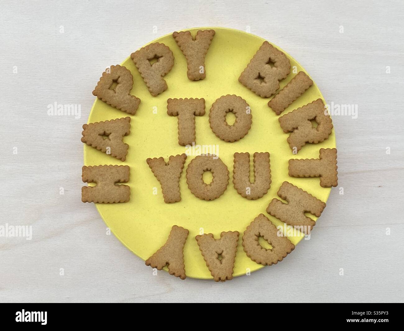 Happy Birthday, kreative Nachricht mit Keksbuchstaben auf einem gelben Teller über einem weißen Holzbrett komponiert - Smartphone-aufgenommenes Stockfoto