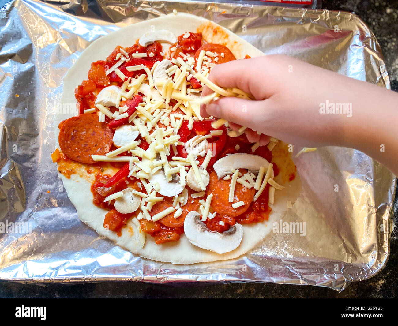 Eine Kinderhand, die geriebenen Käse zu einer hausgemachten Pizza auf einem Backblech mit Blechfolie bedeckt. - Smartphone-aufgenommenes Stockfoto
