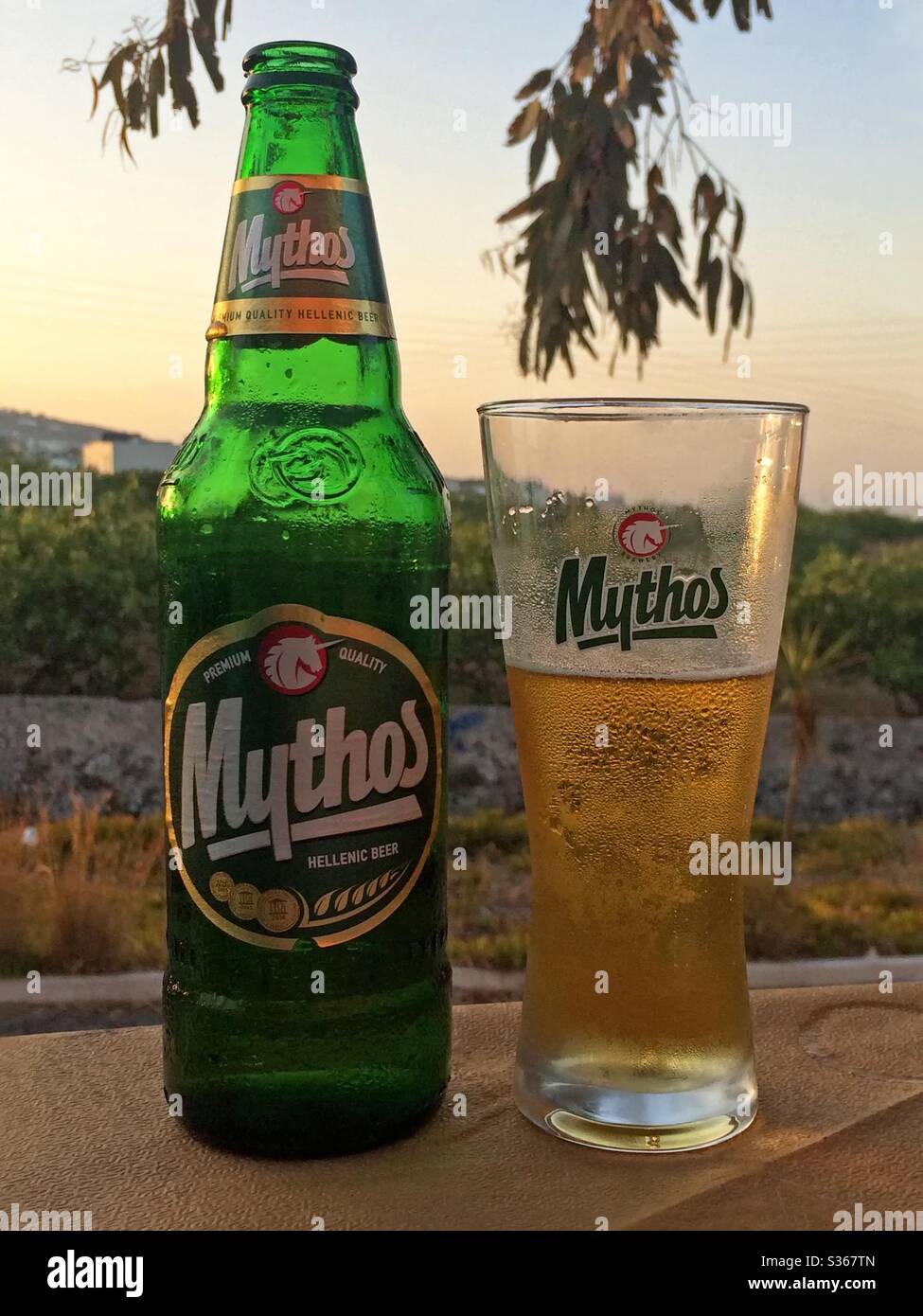 Santorini, Griechenland - 20. Juni 2016: Eine griechische Bierflasche und Glas der Marke Mythos werden am frühen Abend in vertikaler Ansicht auf einem Terrassentisch des Restaurants gezeigt. - Smartphone-aufgenommenes Stockfoto