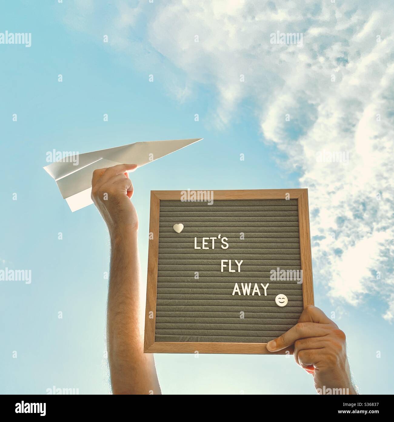 Nahaufnahme eines Mannes, der eine Message Board sagt, lasst uns mit einem Papierflugzeug gegen einen bunten blauen Himmel und verstreute Wolken fliegen. Reise-, Urlaubs- und Fernweh-Konzept - Smartphone-aufgenommenes Stockfoto