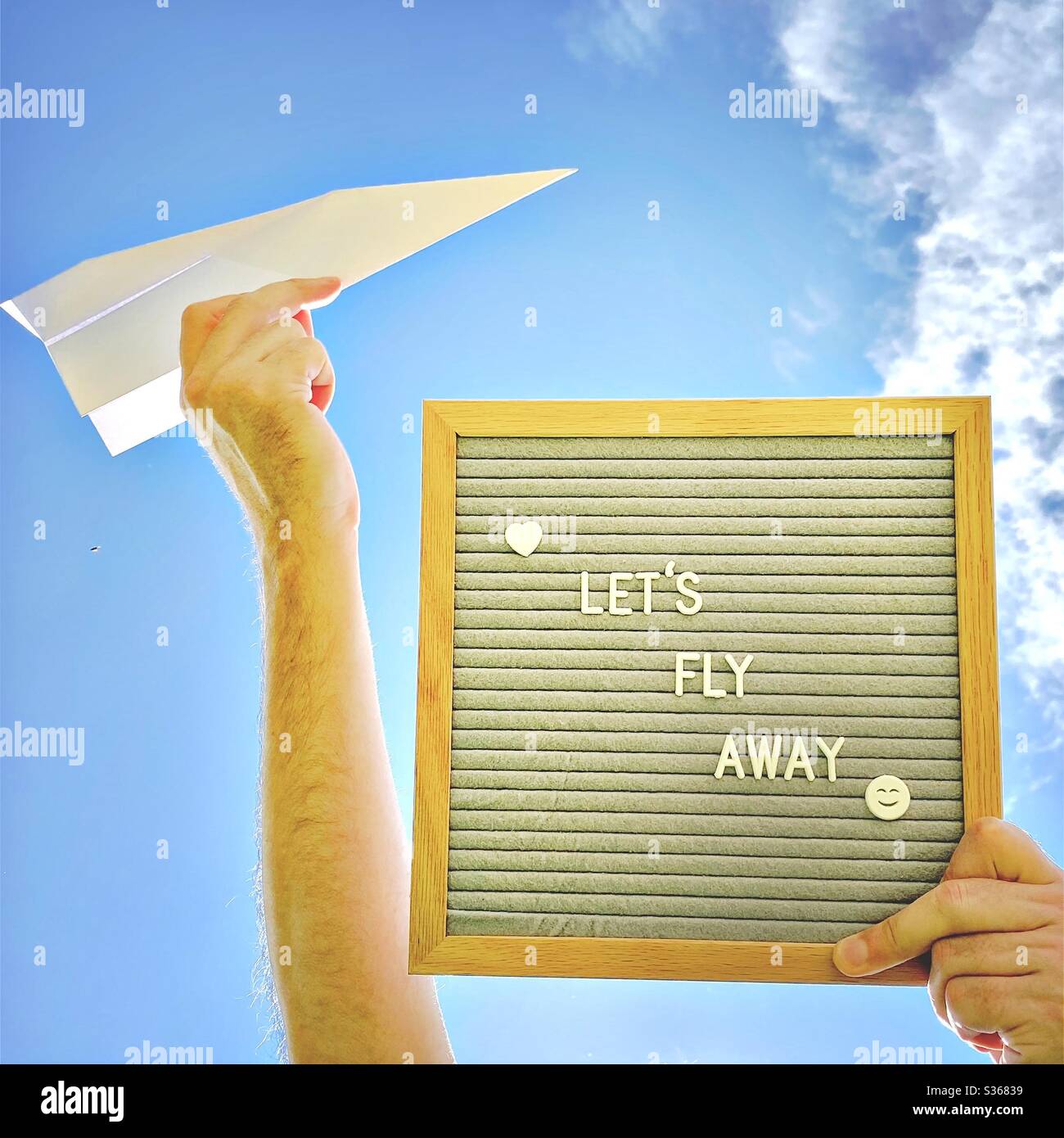 Nahaufnahme eines Mannes, der eine Message Board sagt, lasst uns mit einem Papierflugzeug gegen einen bunten blauen Himmel und verstreute Wolken fliegen. Reise-, Urlaubs- und Fernweh-Konzept - Smartphone-aufgenommenes Stockfoto