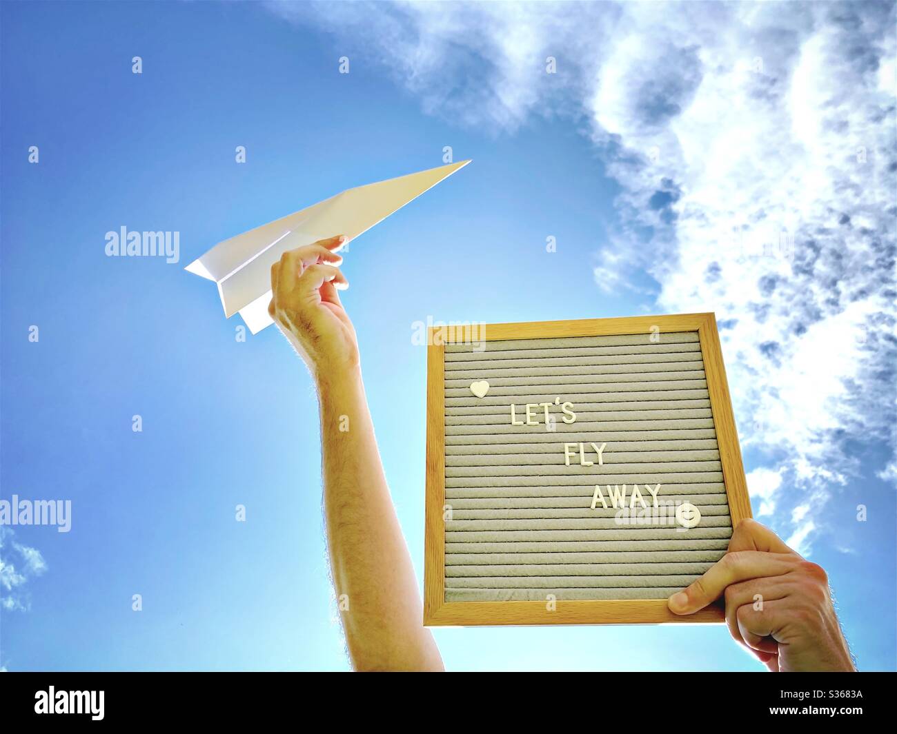 Nahaufnahme eines Mannes, der eine Message Board sagt, lasst uns mit einem Papierflugzeug gegen einen bunten blauen Himmel und verstreute Wolken fliegen. Reise-, Urlaubs- und Fernweh-Konzept - Smartphone-aufgenommenes Stockfoto