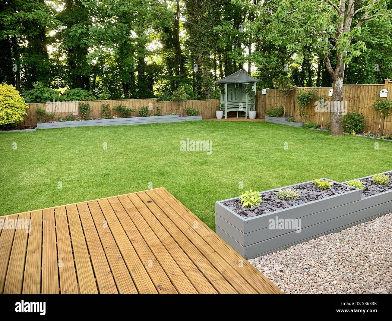 Moderner Garten mit Terrasse, Laube. Fechten, Vogelkisten, Pflanzgefäße und Gras. Umgeben von hohen Bäumen. Stockfoto