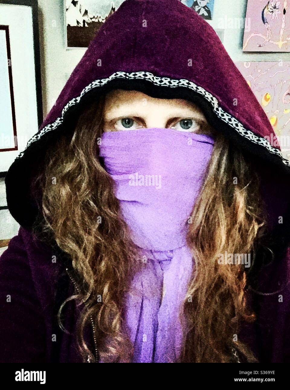 Rothaarige Frau mit blauen Augen, die ein lila Kapuzenpullover und einen lila Schal als COVID19 Maske trägt, die ihr Gesicht bedeckt. Stockfoto