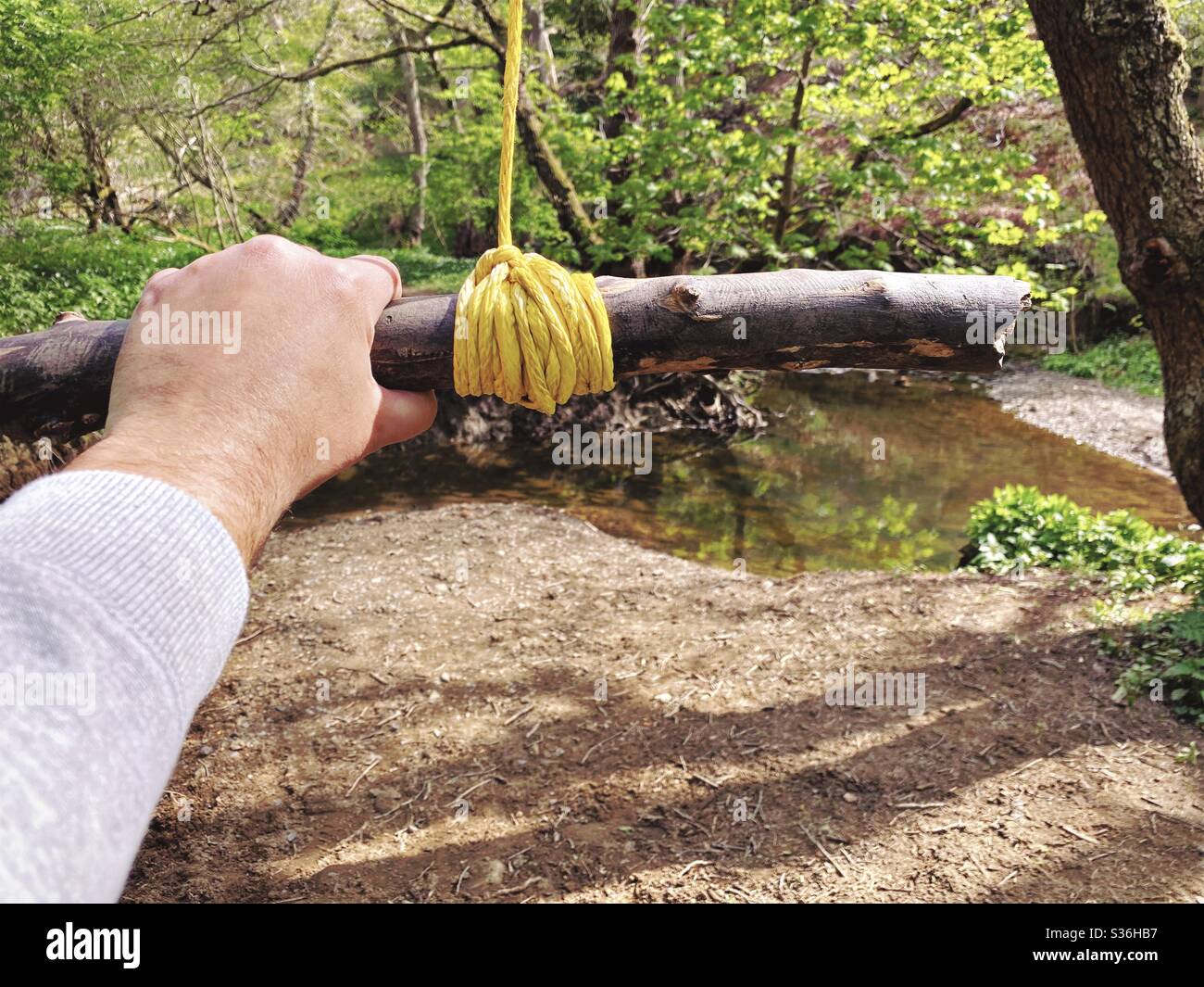 Nahaufnahme von Details eines Mannes, der an einer Seilschaukel hält, die an den Zweigen eines reifen Baumes in einem Wald hängt. Ein kleiner Stab wird mit Knoten als Bügel gebunden, um über Fluß unten zu schwingen - Smartphone-aufgenommenes Stockfoto