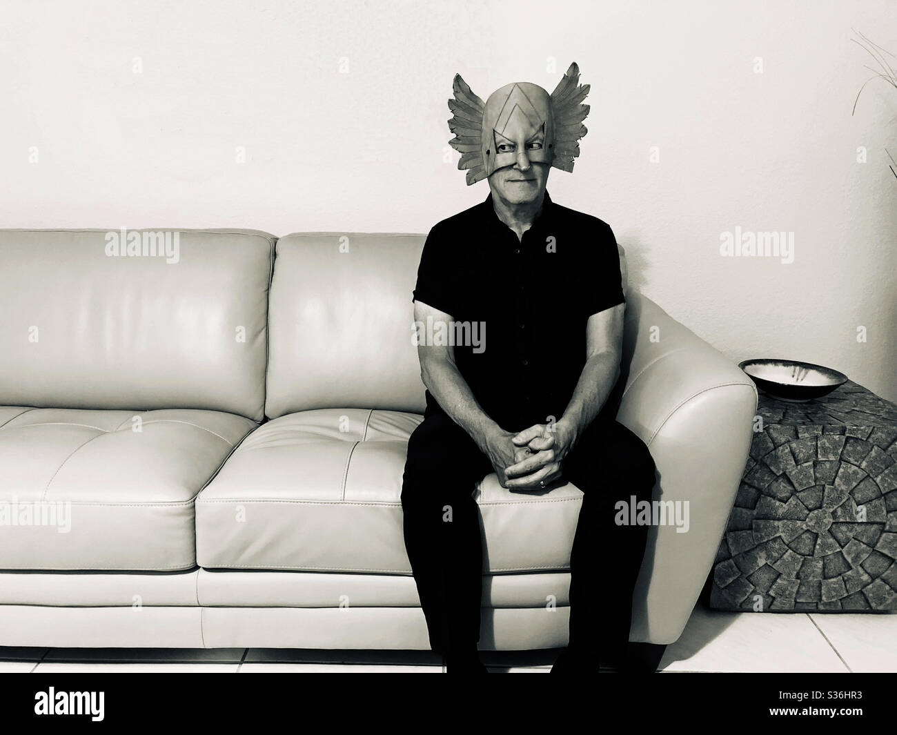 Geheimnisvoll aussehender Mann in einer seltsamen Maske auf einem Sofa sitzend mit seitwärts schauenden Augen. Stockfoto