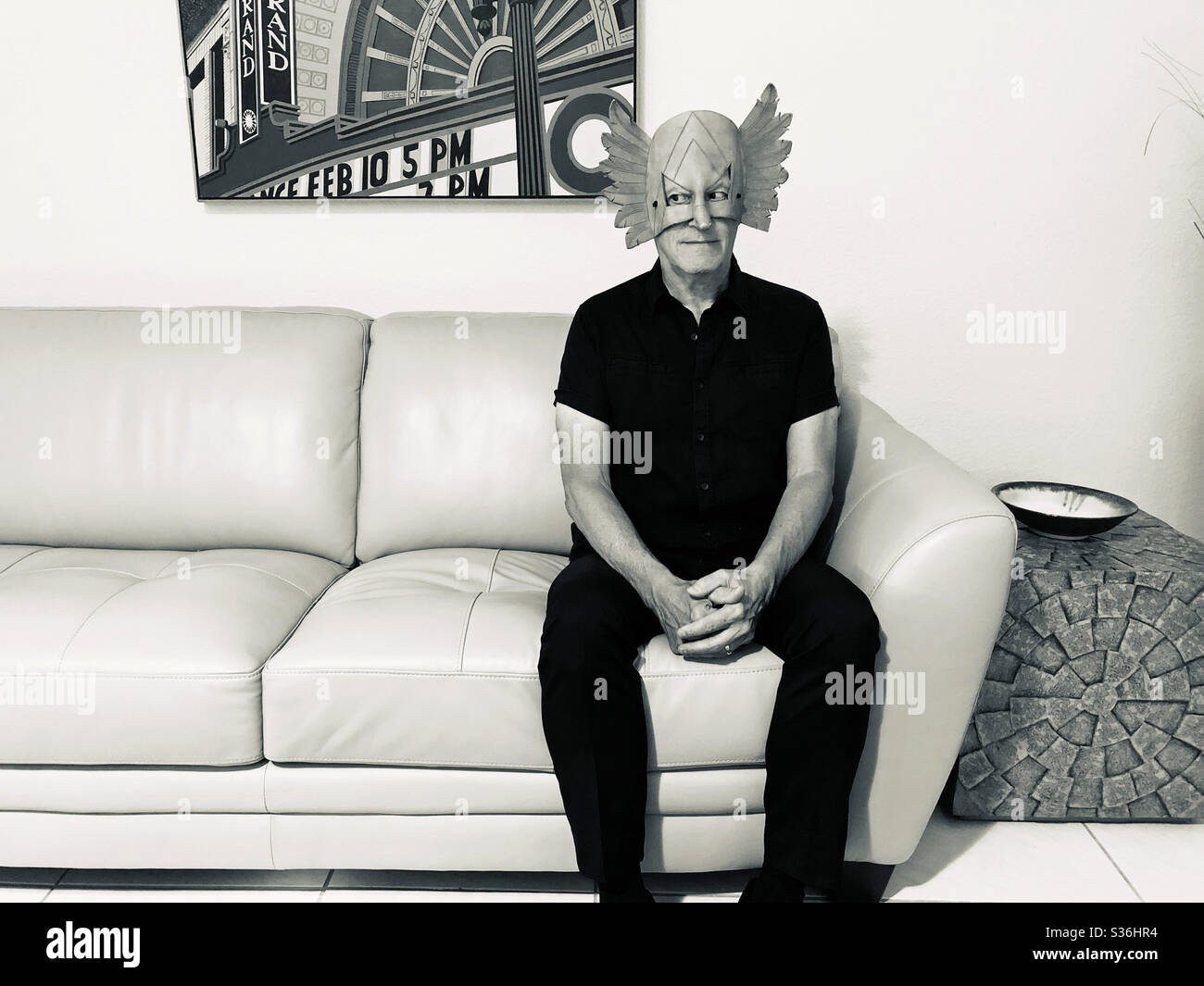 Mann saß auf einem Sofa mit einer seltsamen Maske und sah mit den Augen zur Seite. - Smartphone-aufgenommenes Stockfoto