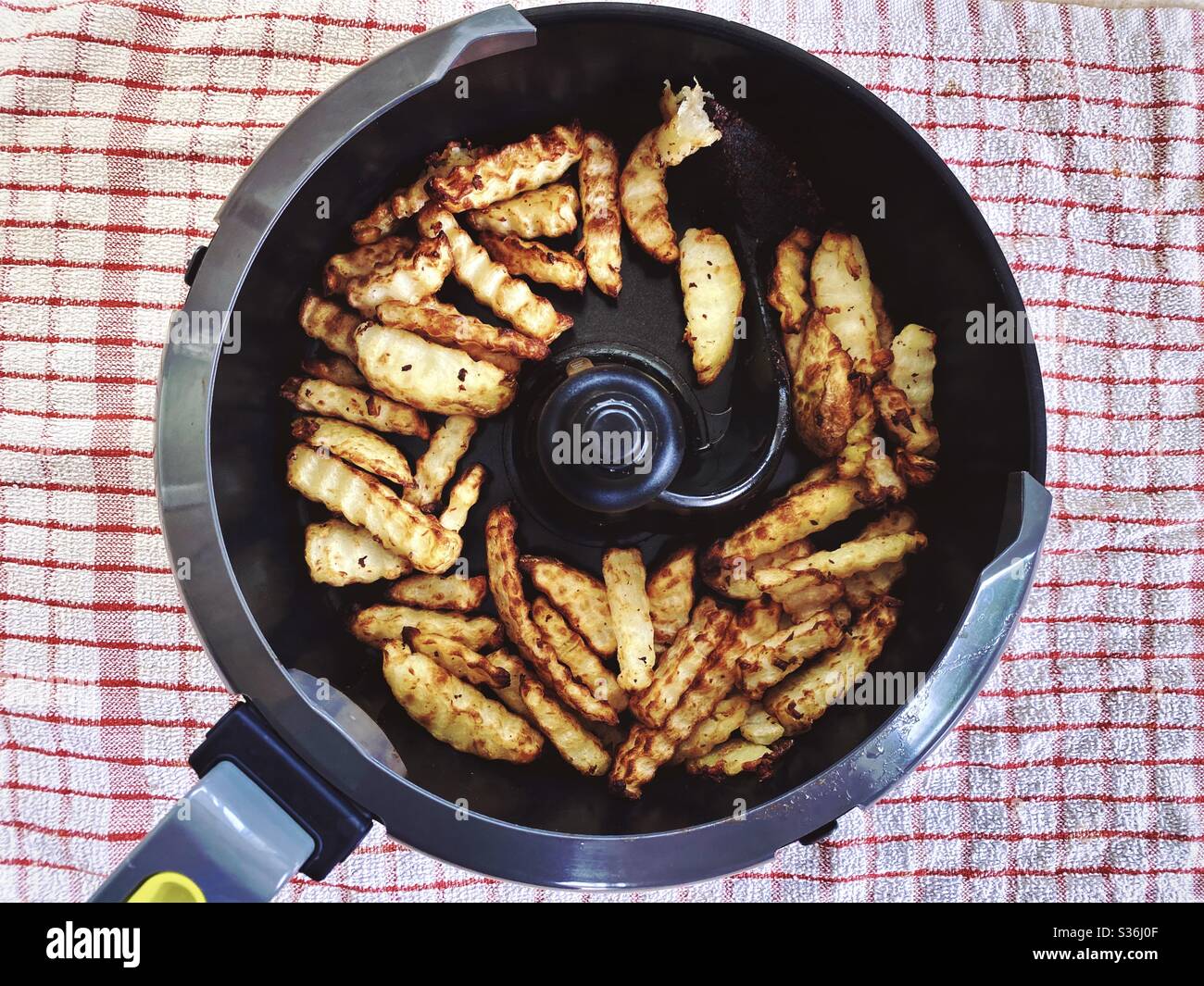 Nahaufnahme der in der Pfanne einer rotierenden Fritteuse geschnittenen Pommes frites. Hausgemachte Pommes frites mit einem in heißer Luft gebratenen Crinkle Cutter, eine gesündere Art zu kochen. Knusprige Kartoffelchips zum Abendessen. - Smartphone-aufgenommenes Stockfoto