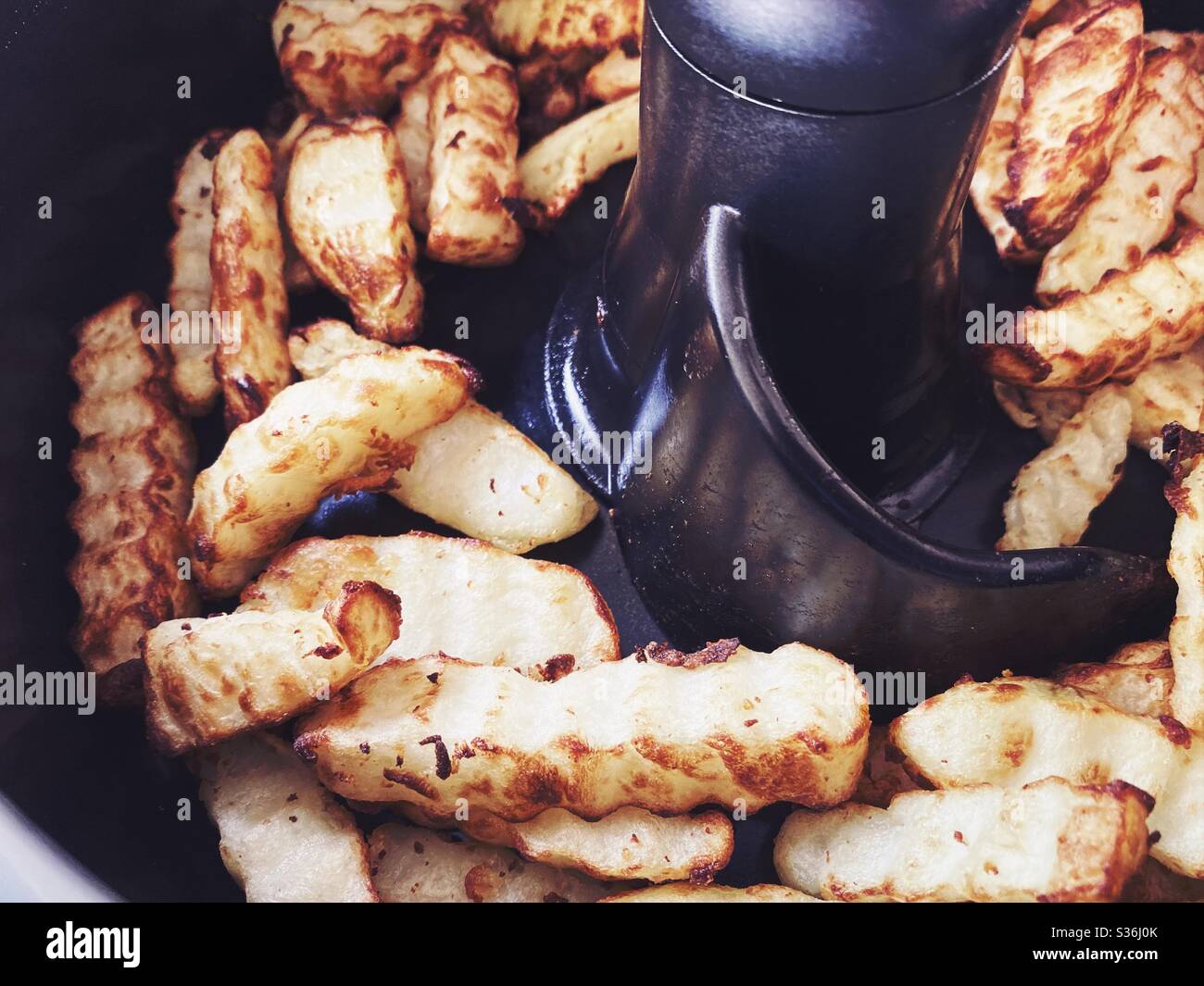 Nahaufnahme der in der Pfanne einer rotierenden Fritteuse geschnittenen Pommes frites. Hausgemachte Pommes frites mit einem in heißer Luft gebratenen Crinkle Cutter, eine gesündere Art zu kochen. Knusprige Kartoffelchips zum Abendessen. - Smartphone-aufgenommenes Stockfoto