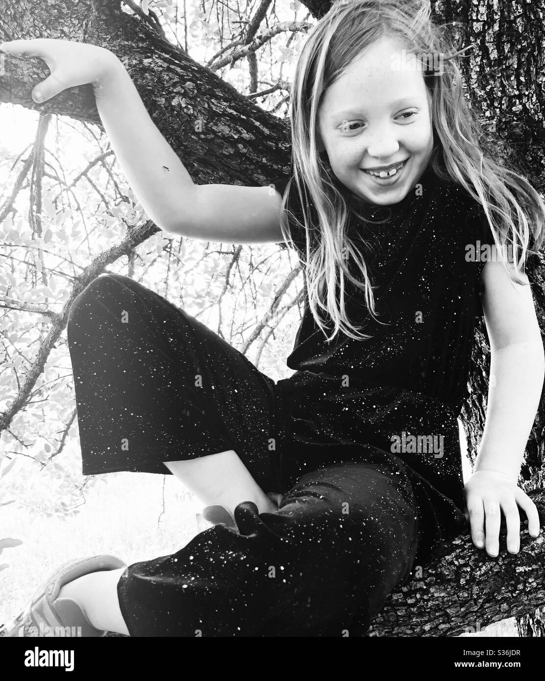 S&W Foto von 7-jähriges Mädchen in schwarzem Overall, das in Ästen eines Baumes sitzt und lächelt Stockfoto