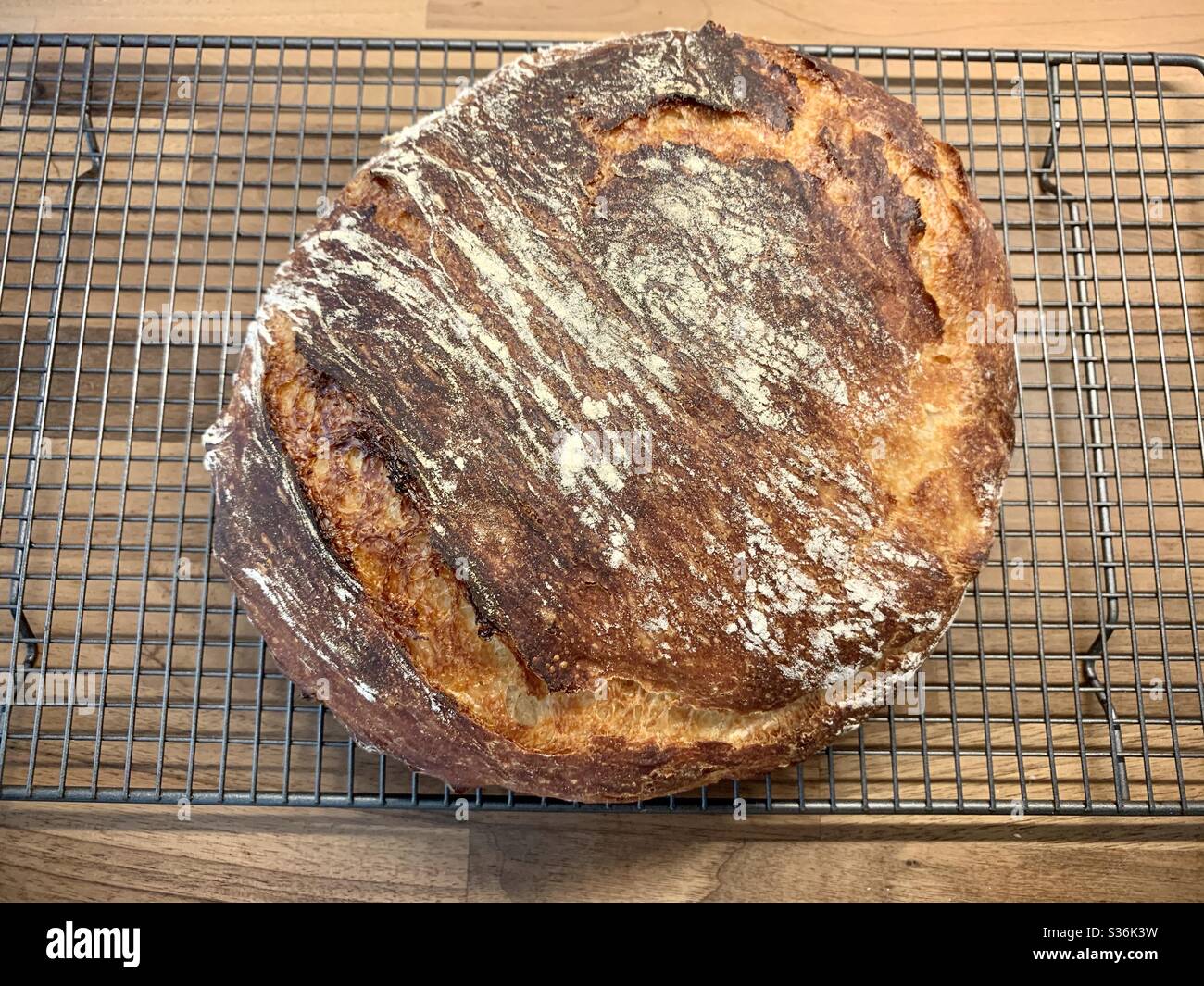 Hausgemachtes Brot. Ein großer knuspriger Laib Brot saß auf einem Drahtgitter auf einer hölzernen Arbeitsfläche. - Smartphone-aufgenommenes Stockfoto