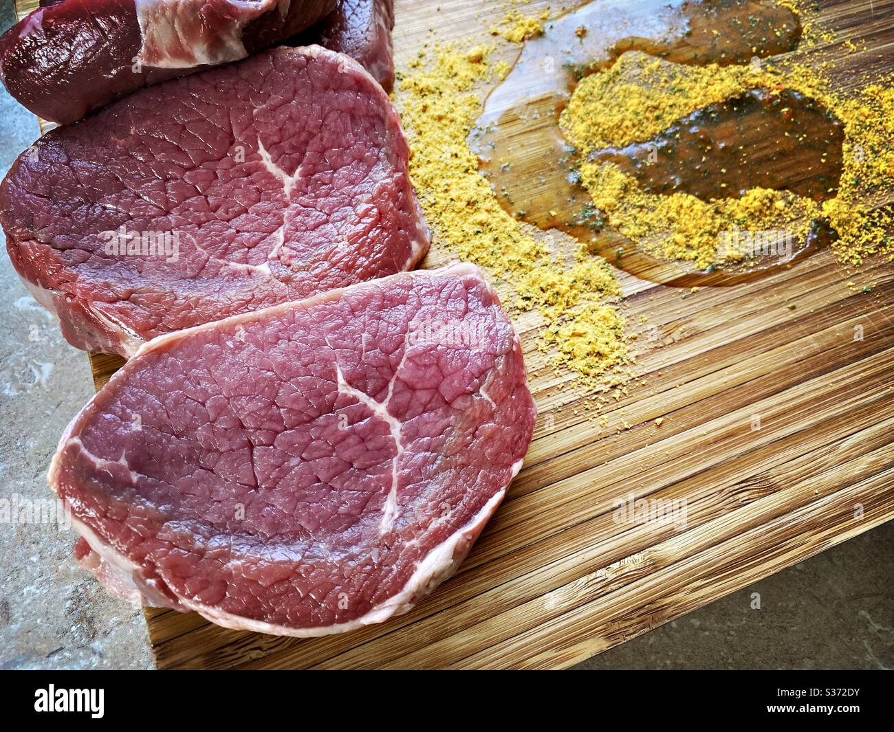 Nahaufnahme von rohen Rinderhacksteaks auf einem rustikalen Holzbrett mit Knoblauchgewürzen und Olivenöl. Marinieren von Fleisch in der Küche vor dem Kochen Abendessen. - Smartphone-aufgenommenes Stockfoto