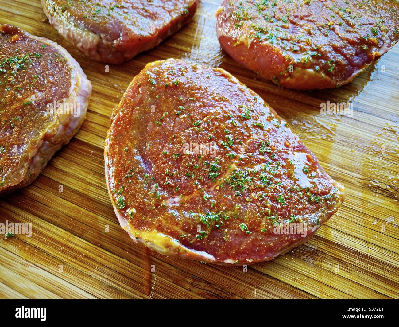 Nahaufnahme von rohen Rindssteaks, mariniert mit Knoblauch, Olivenöl und Gewürzen. Zubereitung von Zutaten für das Abendessen in der Küche auf einer rustikalen Holztheke - Smartphone-aufgenommenes Stockfoto