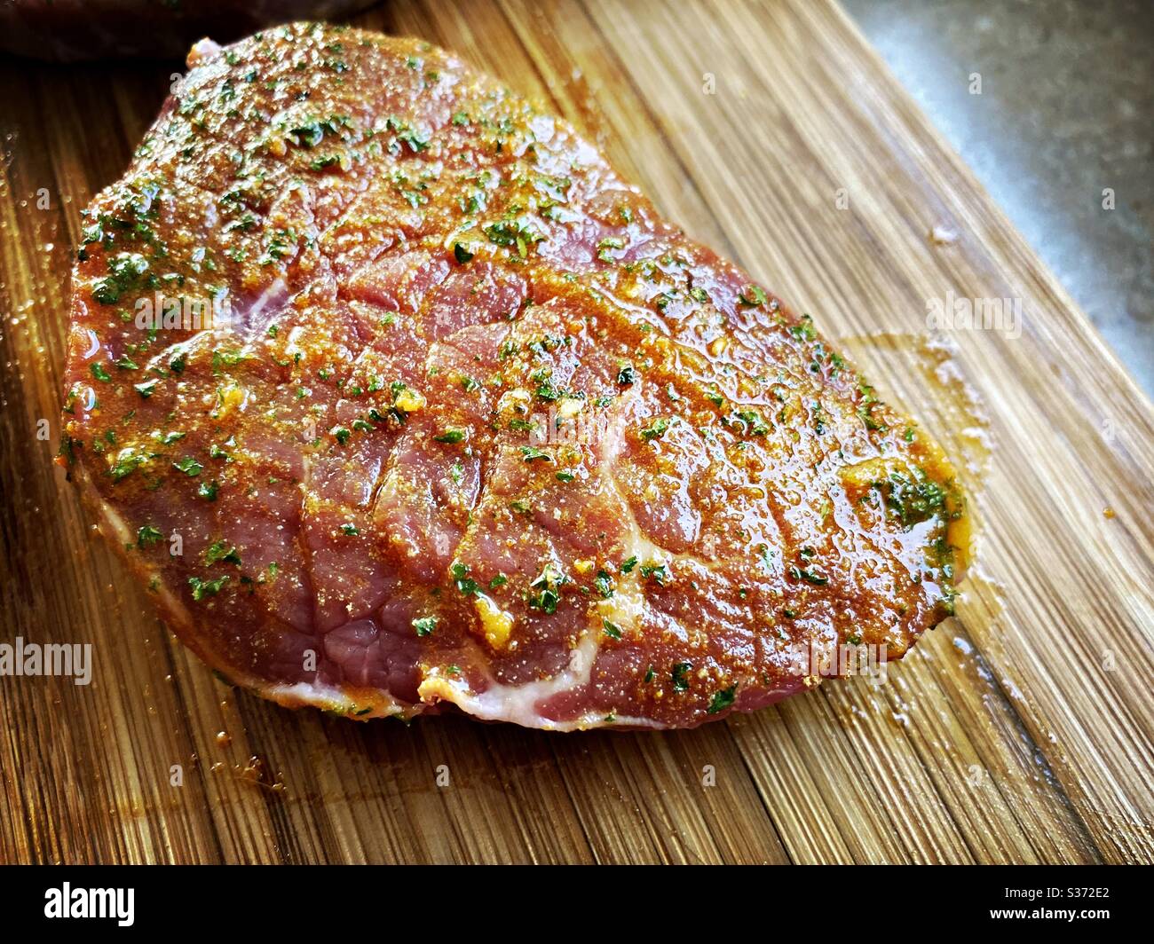 Nahaufnahme von rohen Rindssteaks, mariniert mit Knoblauch, Olivenöl und Gewürzen. Zubereitung von Zutaten für das Abendessen in der Küche auf einer rustikalen Holztheke - Smartphone-aufgenommenes Stockfoto