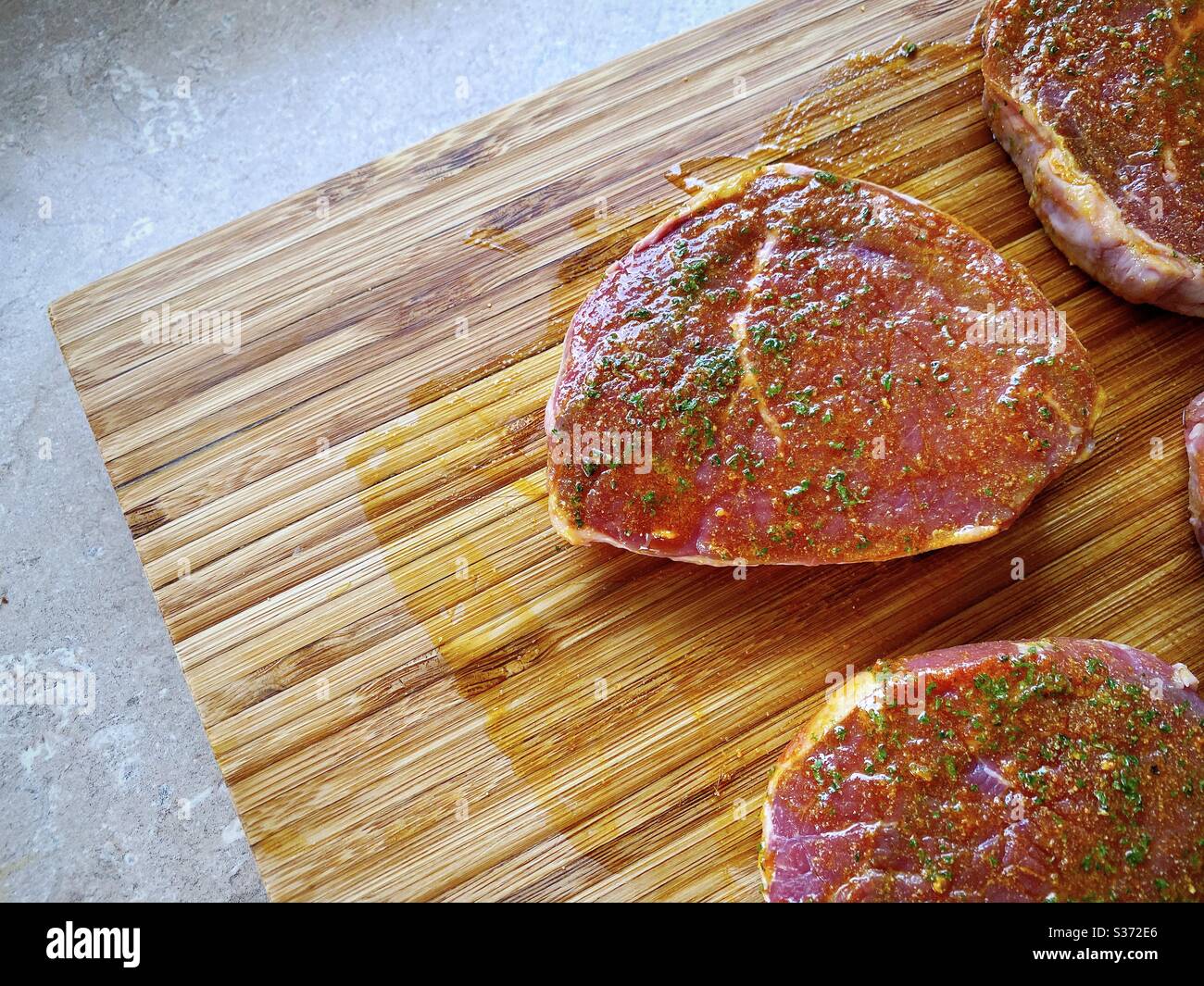 Nahaufnahme von rohen Rindssteaks, mariniert mit Knoblauch, Olivenöl und Gewürzen. Zubereitung von Zutaten für das Abendessen in der Küche auf einer rustikalen Holztheke - Smartphone-aufgenommenes Stockfoto
