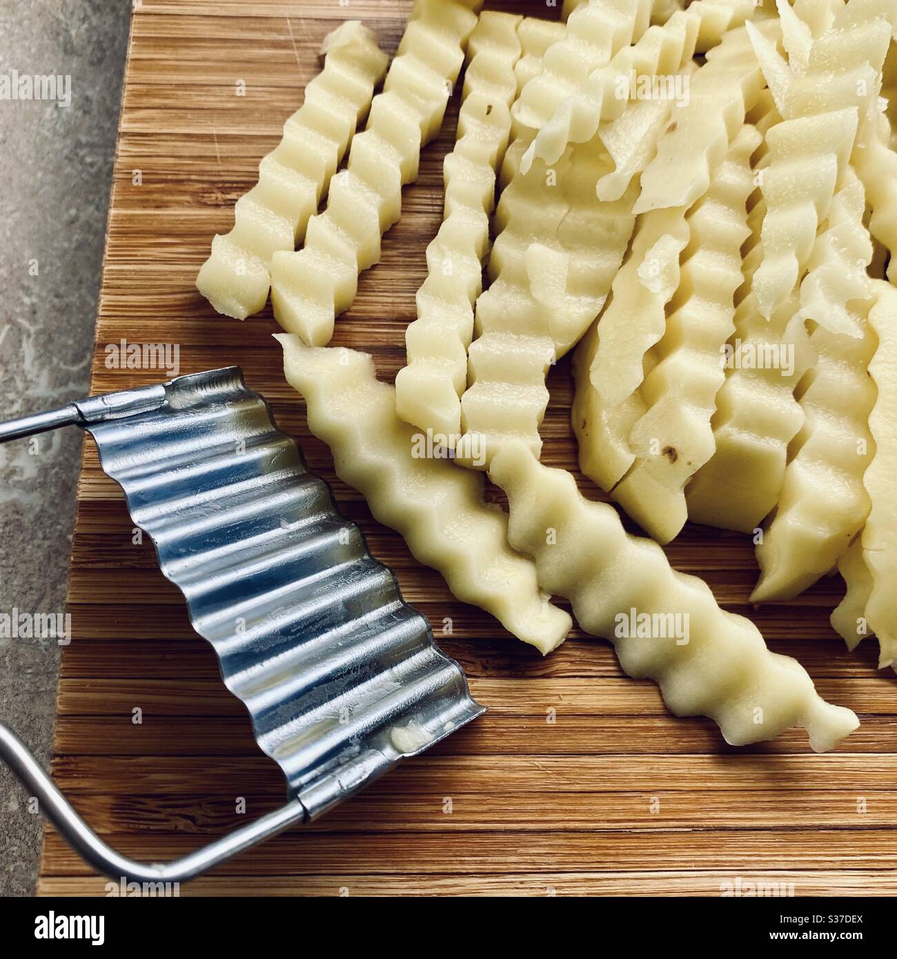 Zubereitung von Crinkle Cut Pommes (US) oder Pommes (UK) in der Küche auf einem rustikalen Holzschneidebrett. Mit einer Crinkle Cut Stahlklinge Kartoffeln zum Braten fertig hacken. - Smartphone-aufgenommenes Stockfoto