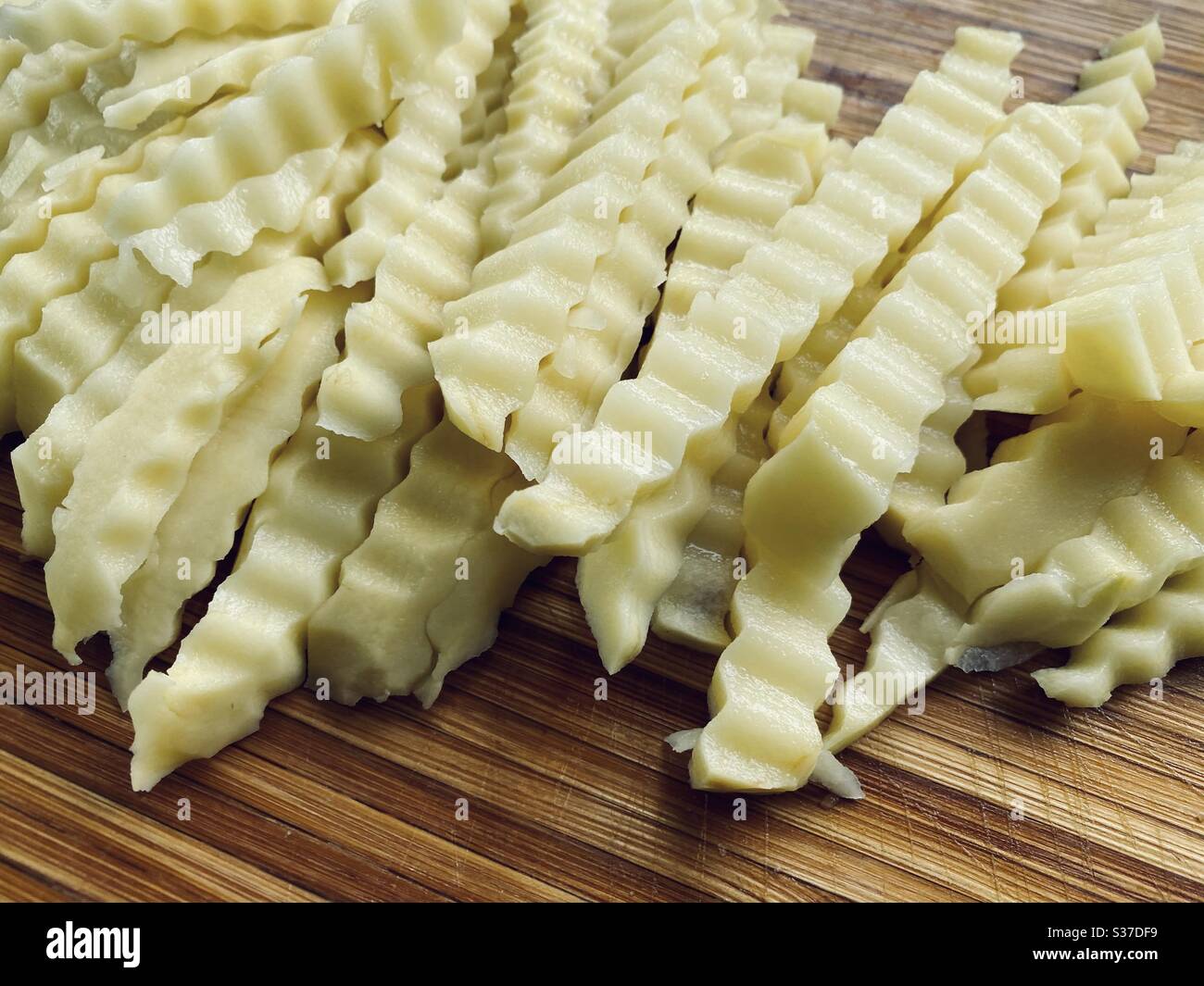 Zubereitung von Crinkle Cut Pommes (US) oder Pommes (UK) in der Küche auf einem rustikalen Holzschneidebrett. Kartoffelscheiben mit zerklüfteten Rändern zum Braten bereit. Stockfoto