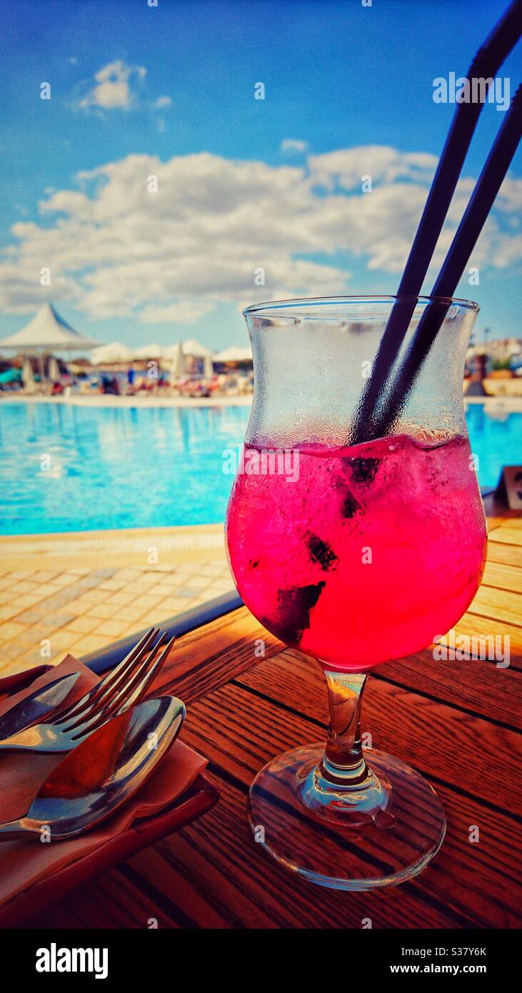 Ein Foto eines schönen, lebendigen roten Cocktailgetränks mit Strohhalmen und Besteck, vor einem türkisfarbenen Pool an einem sonnigen Tag. Sommerurlaub am Strand. Stockfoto