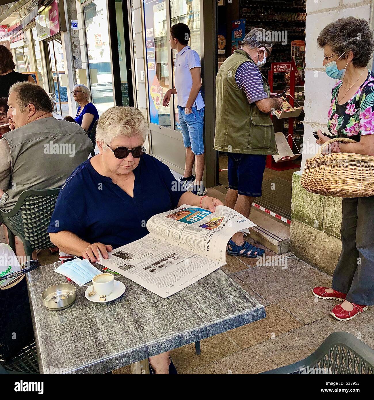 Ältere Frau beim Lesen einer Zeitung an einem Café-Tisch - Frankreich. - Smartphone-aufgenommenes Stockfoto