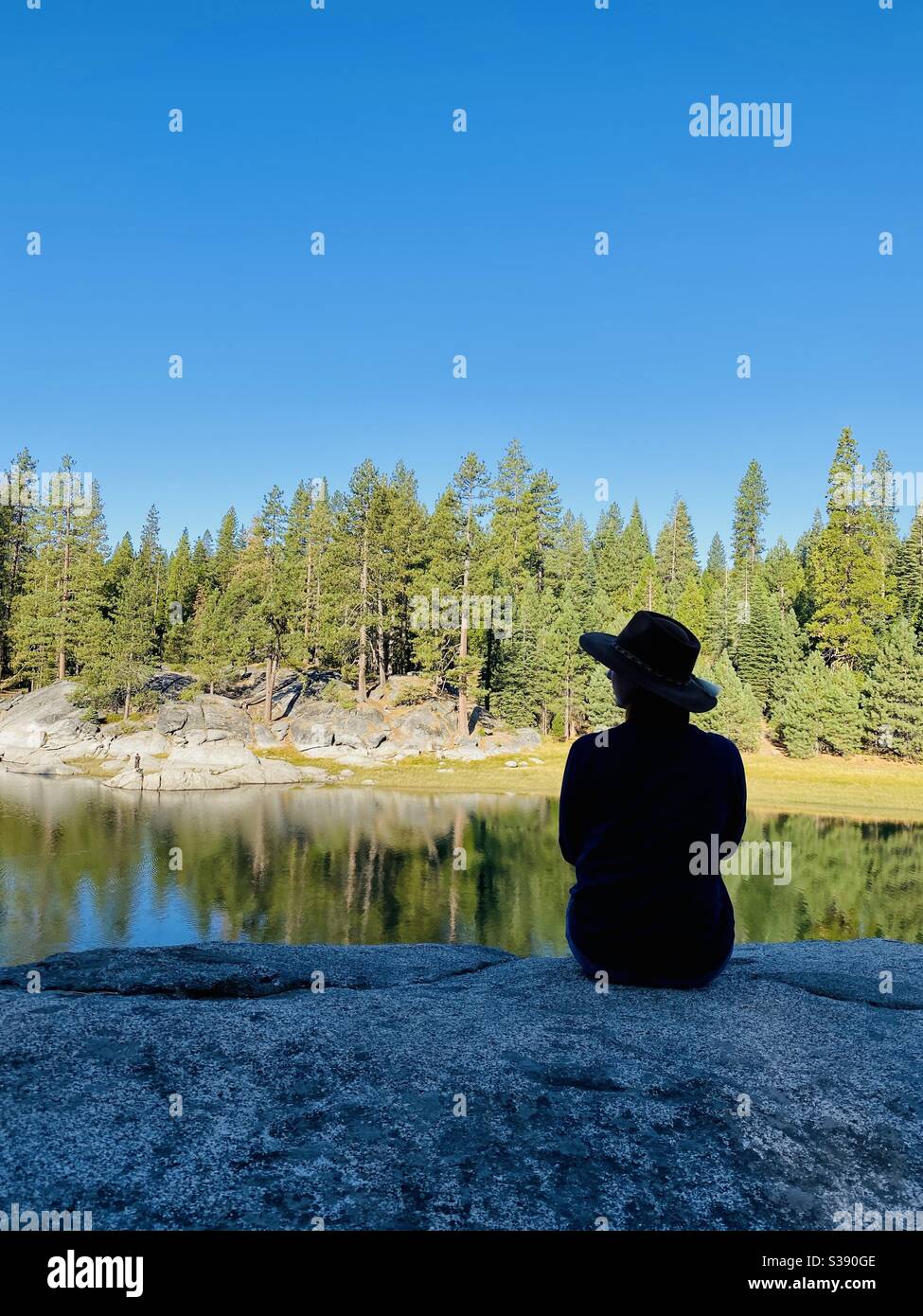 Eine Frau sitzt allein und blickt auf den See und den Wald. Shaver Lake, Kalifornien, USA. - Smartphone-aufgenommenes Stockfoto