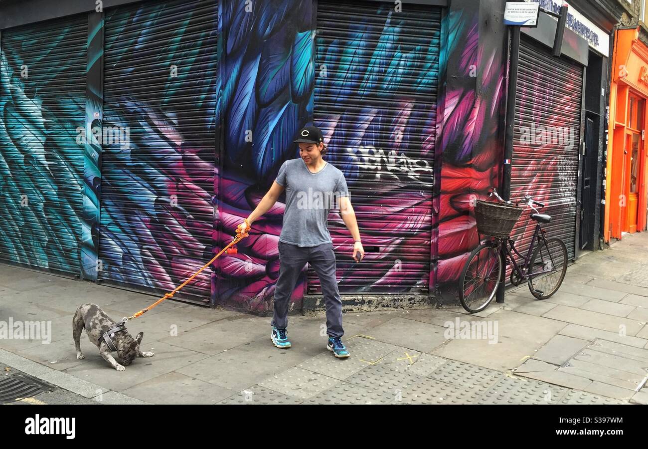 Mann, der vor der französischen Bulldogge an der Leine läuft Graffiti bemalte Schaufelläden in der Nähe der Brick Lane London - Smartphone-aufgenommenes Stockfoto