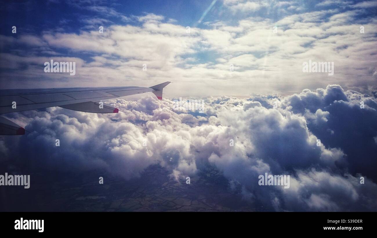 Ein Foto, das aus dem Inneren eines Flugzeugs auf den Flügel des Flugzeugs blickt, Blick über flauschige weiße Wolken und bewohntes Land darunter. Stockfoto
