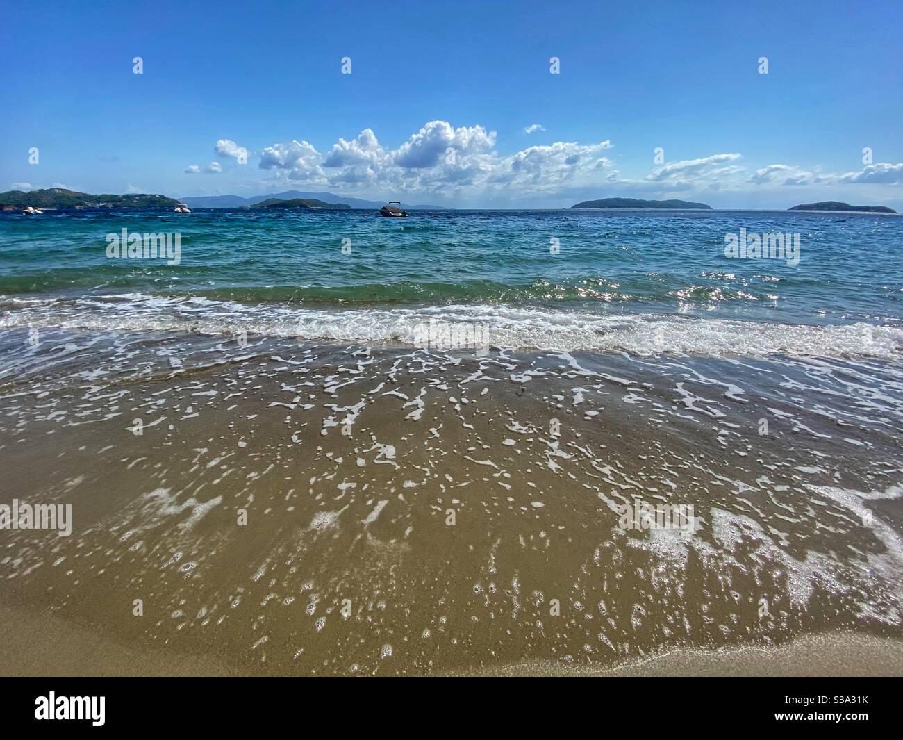 Meereslandschaft mit Schaum auf dem Sand, Booten und kleinen grünen Inseln auf der Insel Skiathos in Griechenland. - Smartphone-aufgenommenes Stockfoto