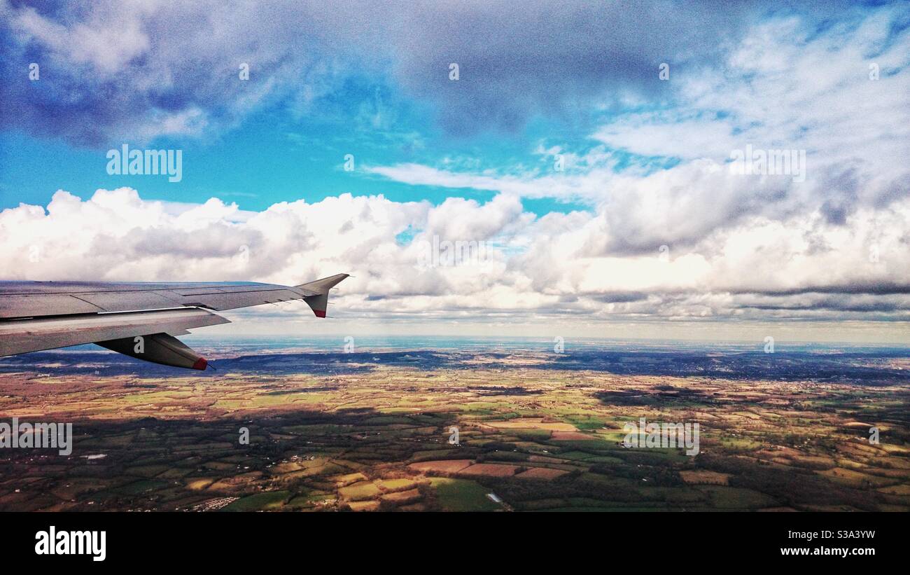 Ein Foto, das aus einem Flugzeugfenster über Land blickt. Horizont, Flugzeug Flügel, Land unten und flauschige weiße Wolken. Stockfoto