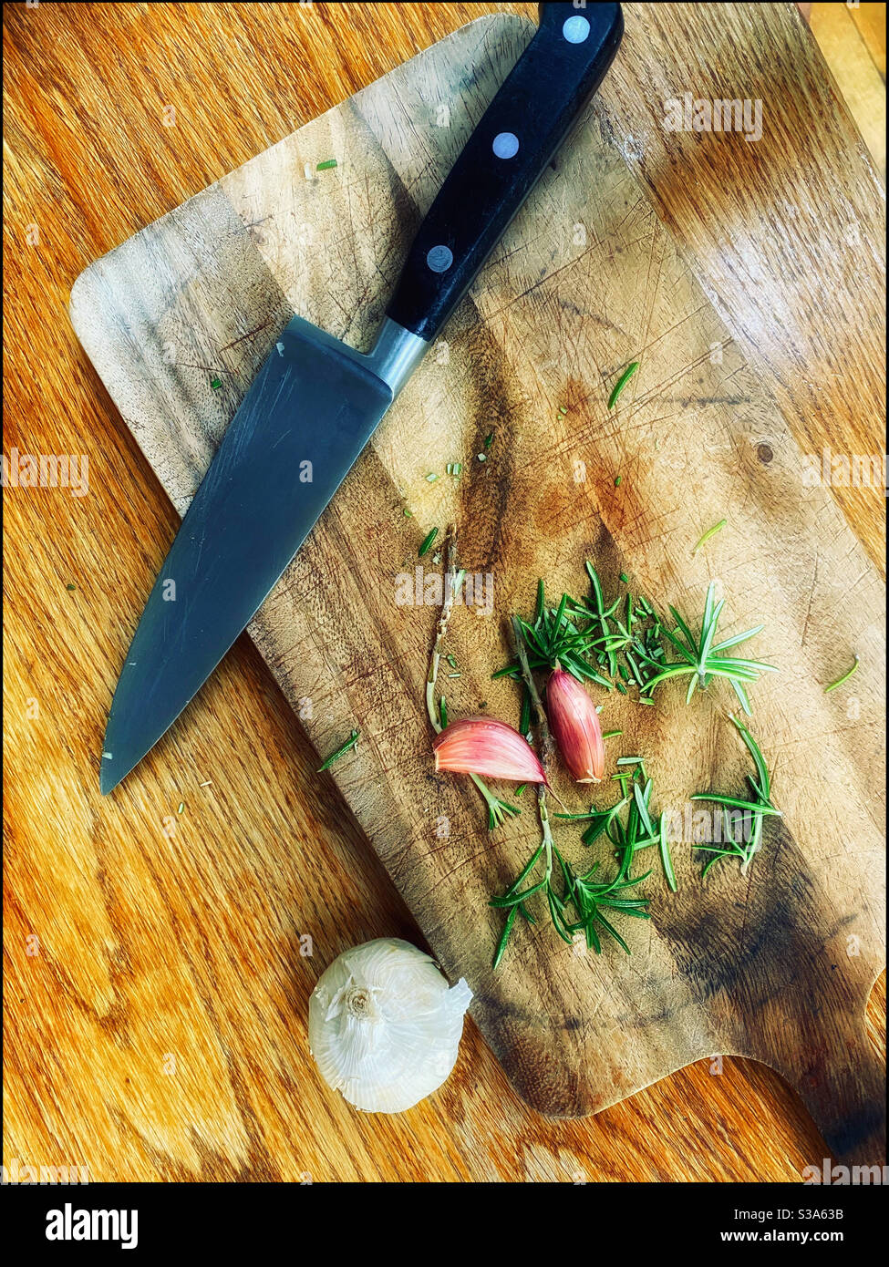 Ein Blick von oben auf ein Schneidebrett mit Rosmarin, Knoblauch und einem Küchenmesser. Aromen werden vorbereitet! Bildnachweis - ©️ COLIN HOSKINS. - Smartphone-aufgenommenes Stockfoto