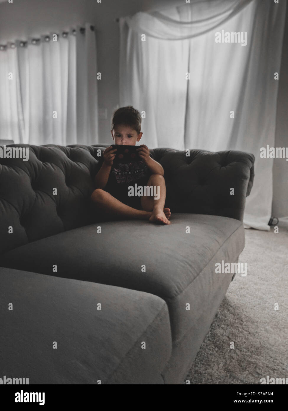 Generation Alpha, ein Junge, schon in jungen Jahren technologiesüchtig. Sitzen auf einer grauen Couch im Wohnzimmer, im Dunkeln, fasziniert vom Bildschirm. Stockfoto