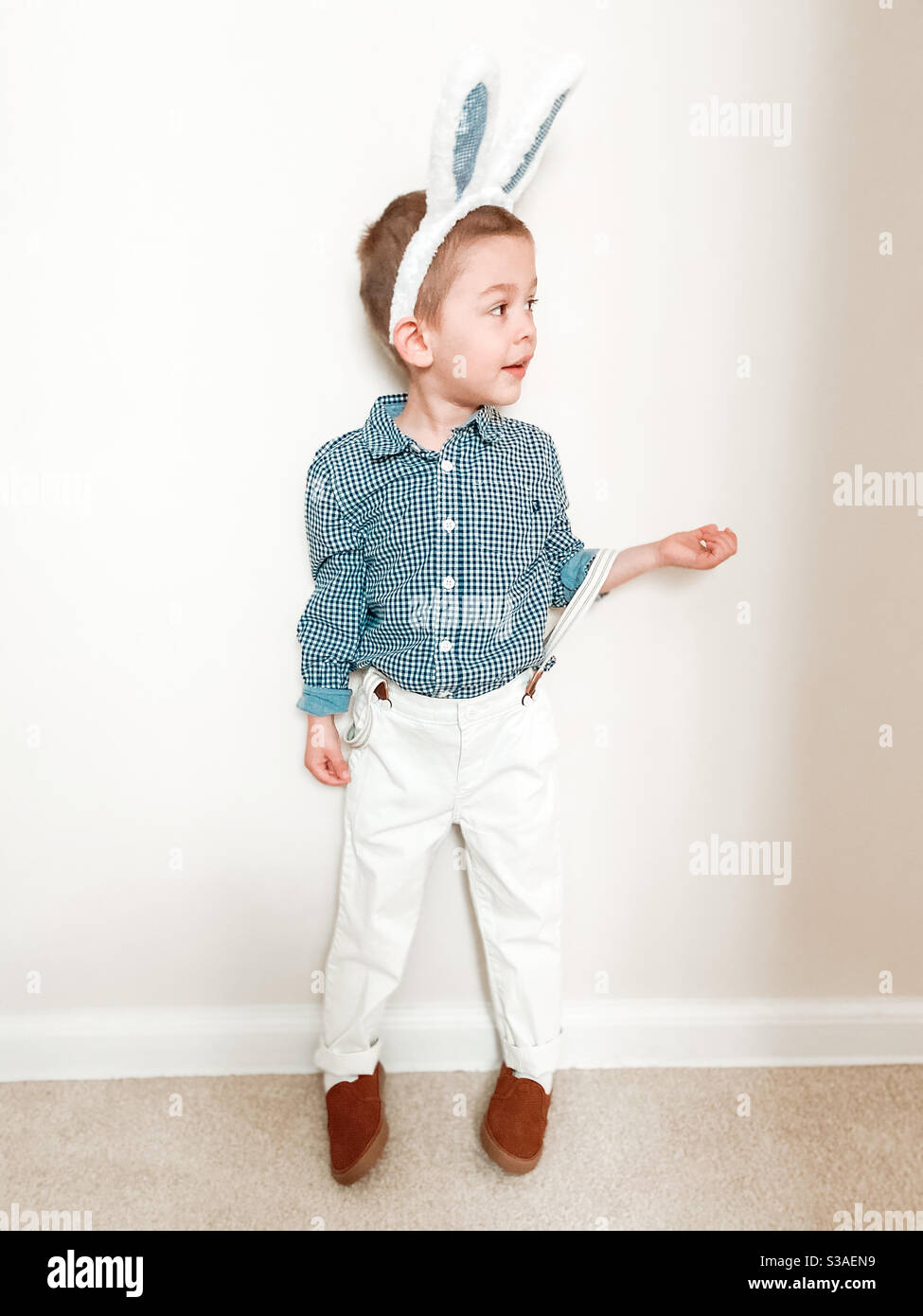 Der kleine blonde Junge feiert Ostern mit Hasenohren, weißen Hosen und einem karierten Hemd. Stockfoto