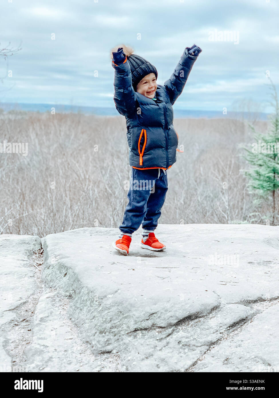 Der vierjährige klettert erfolgreich den Berg hinauf, um ein Foto mit einer schönen Aussicht zu machen. Er hat seine Hände hoch, fröhlich und lächelnd. Stockfoto