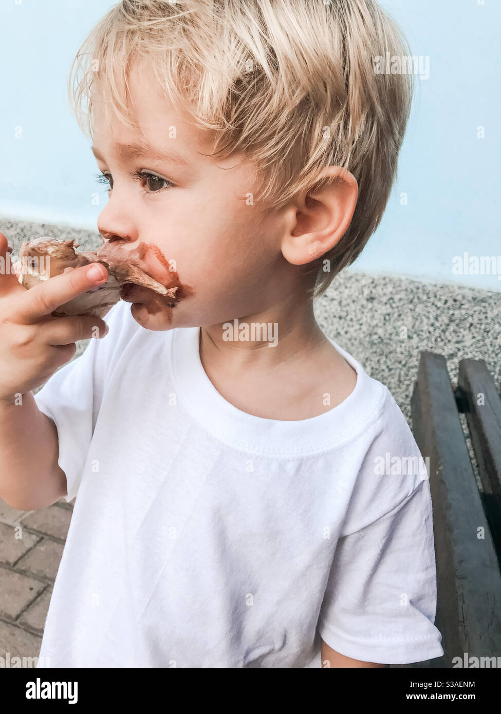 2 Jahre alt blonde junge essen Schokolade Eis mit Ein weißes T-Shirt auf der Außenseite auf einem heißen und Sonniger Tag Stockfoto