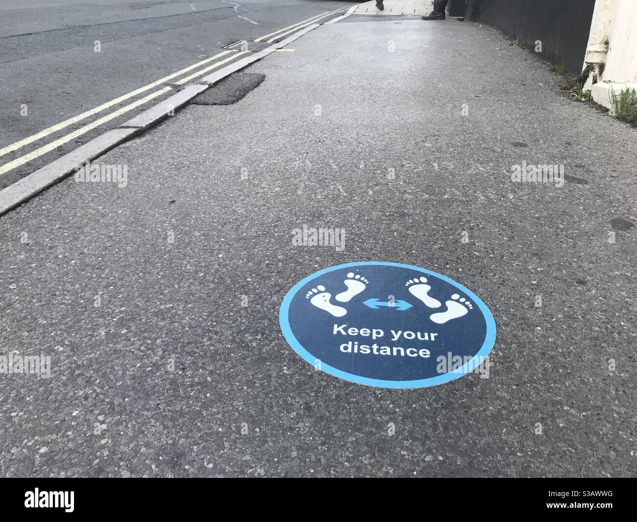 Ein Schild auf Bürgersteig sagt halten Sie Ihren Abstand. Die Symbole der Fußabdrücke sind sichtbar und die Straße führt in die Ferne Stockfoto