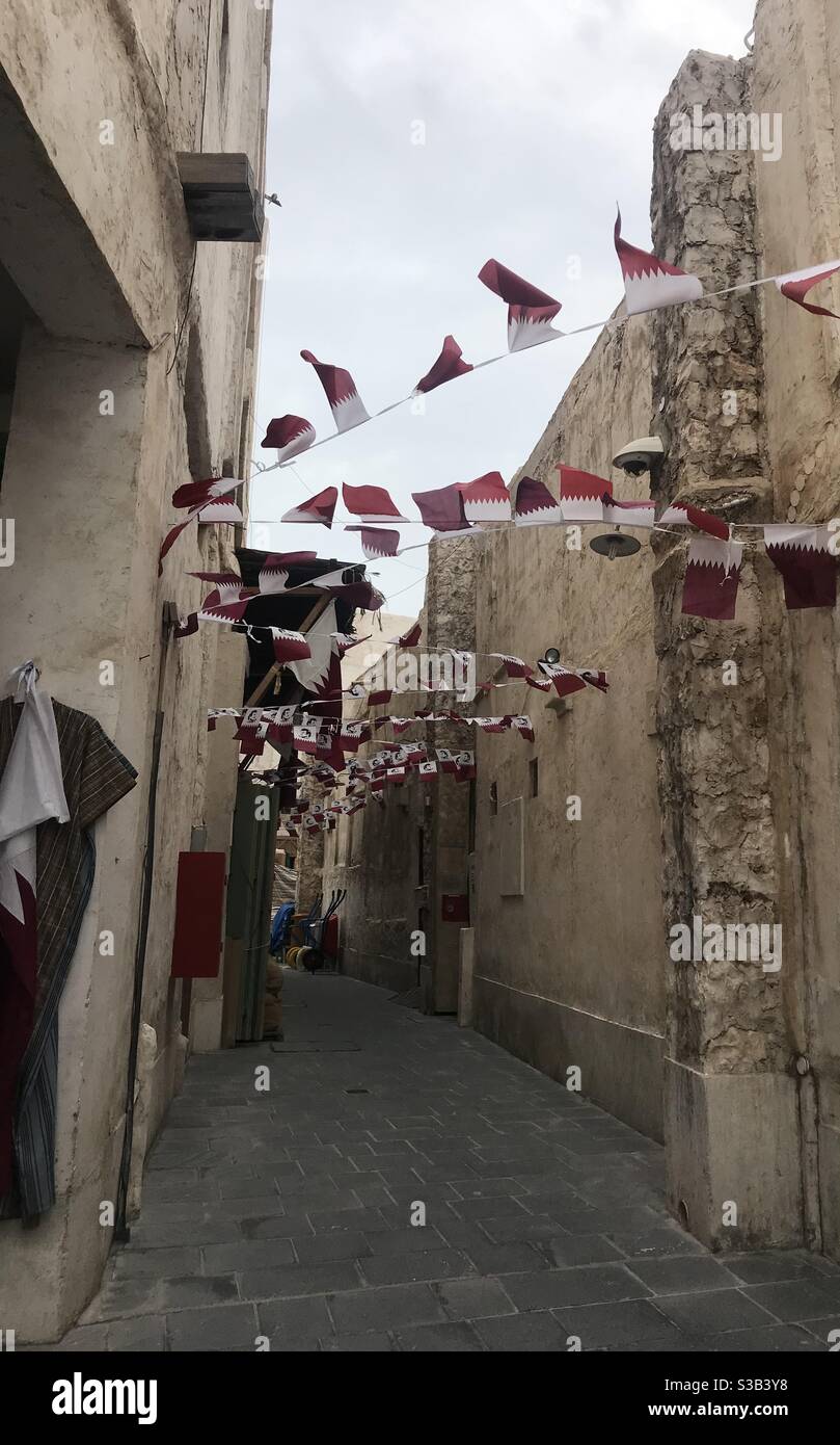 Kette von katarischen Flaggen, die an einem windigen Tag in Souq Waqif, Doha, Katar, zwischen Gebäuden hängen - Smartphone-aufgenommenes Stockfoto