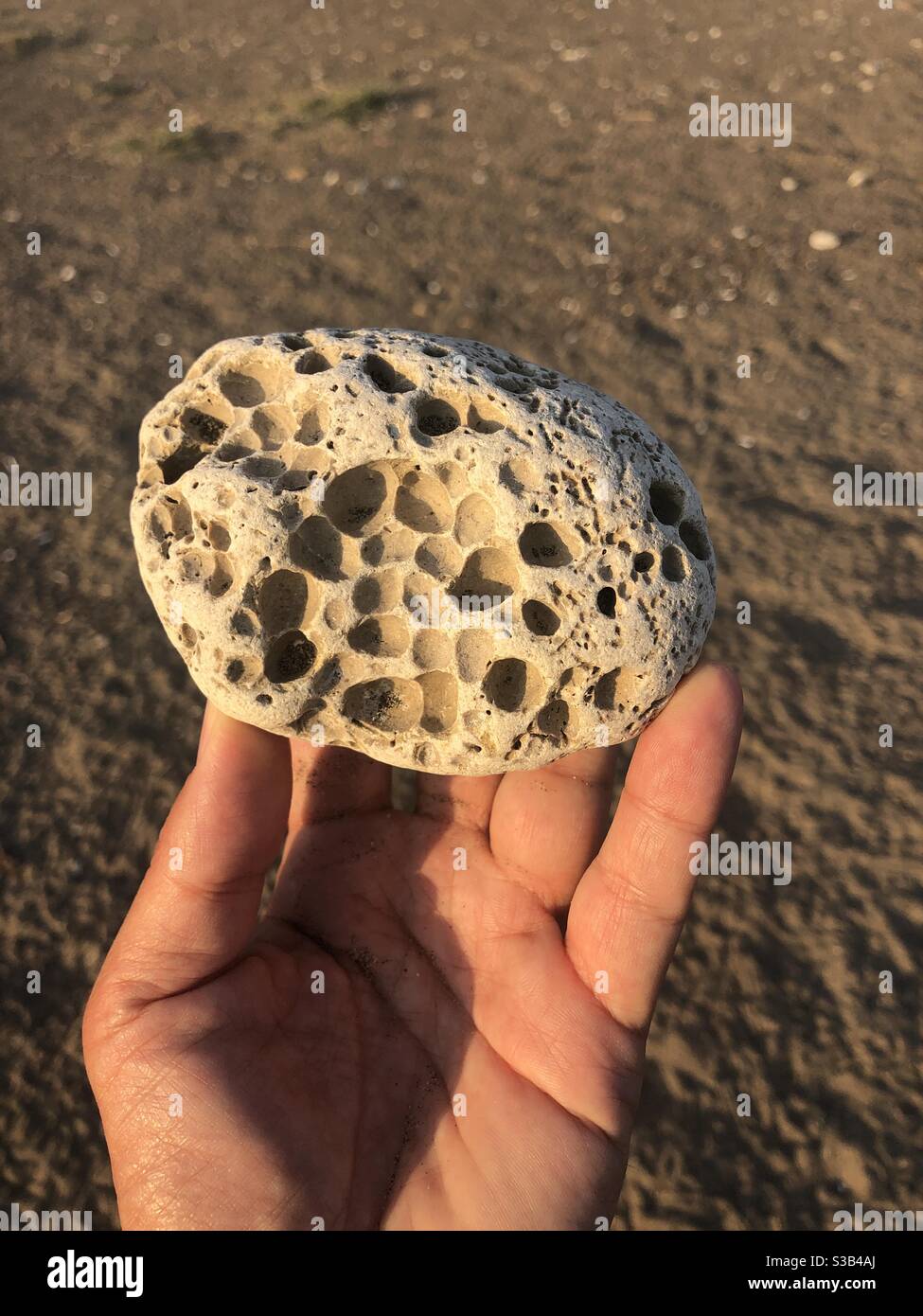 Eine Person, die einen strukturierten Kieselstein in der Hand gegen Sand hält Hintergrund - Smartphone-aufgenommenes Stockfoto